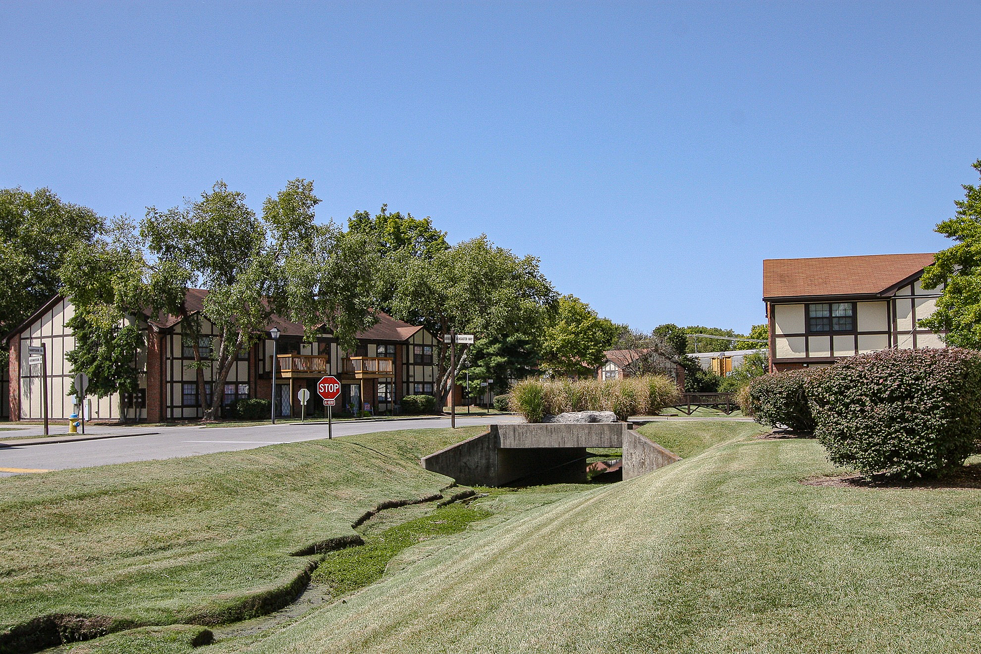 Charming residential area featuring Tudor-style apartments surrounded by lush greenery and a quaint bridge.