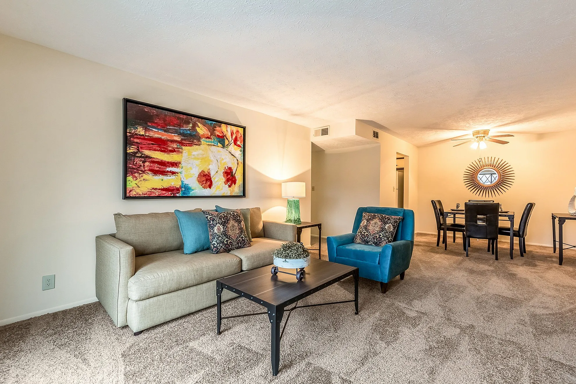 Elegant living room with plush carpeting, featuring a cozy beige sofa, vibrant artwork, and a stylish dining area in the background.