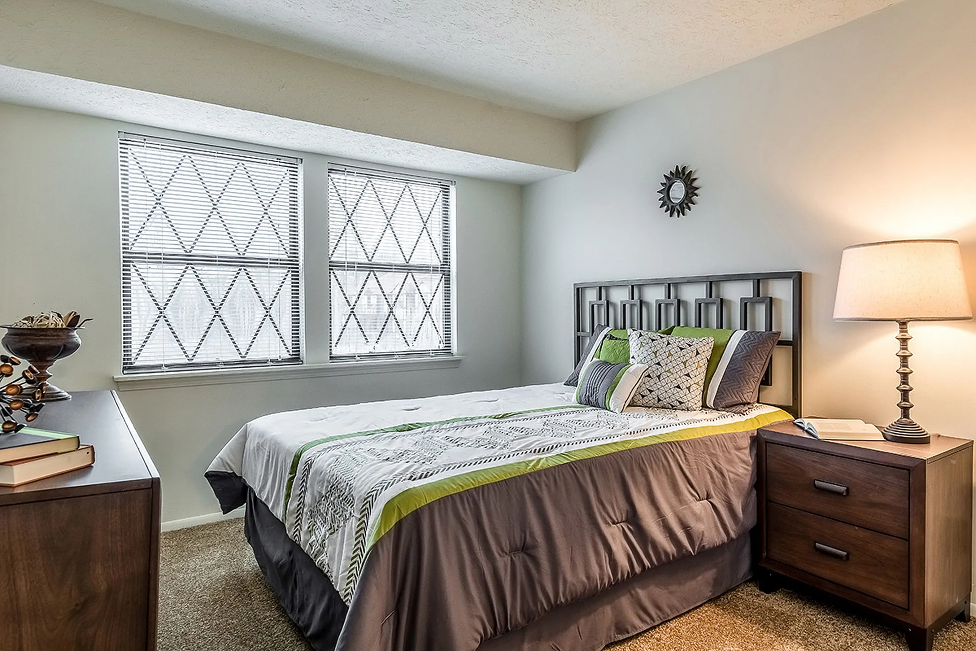 Inviting bedroom with stylish decor, featuring a cozy bed adorned with elegant pillows, large windows with patterned blinds, and a warm table lamp beside a wooden nightstand.