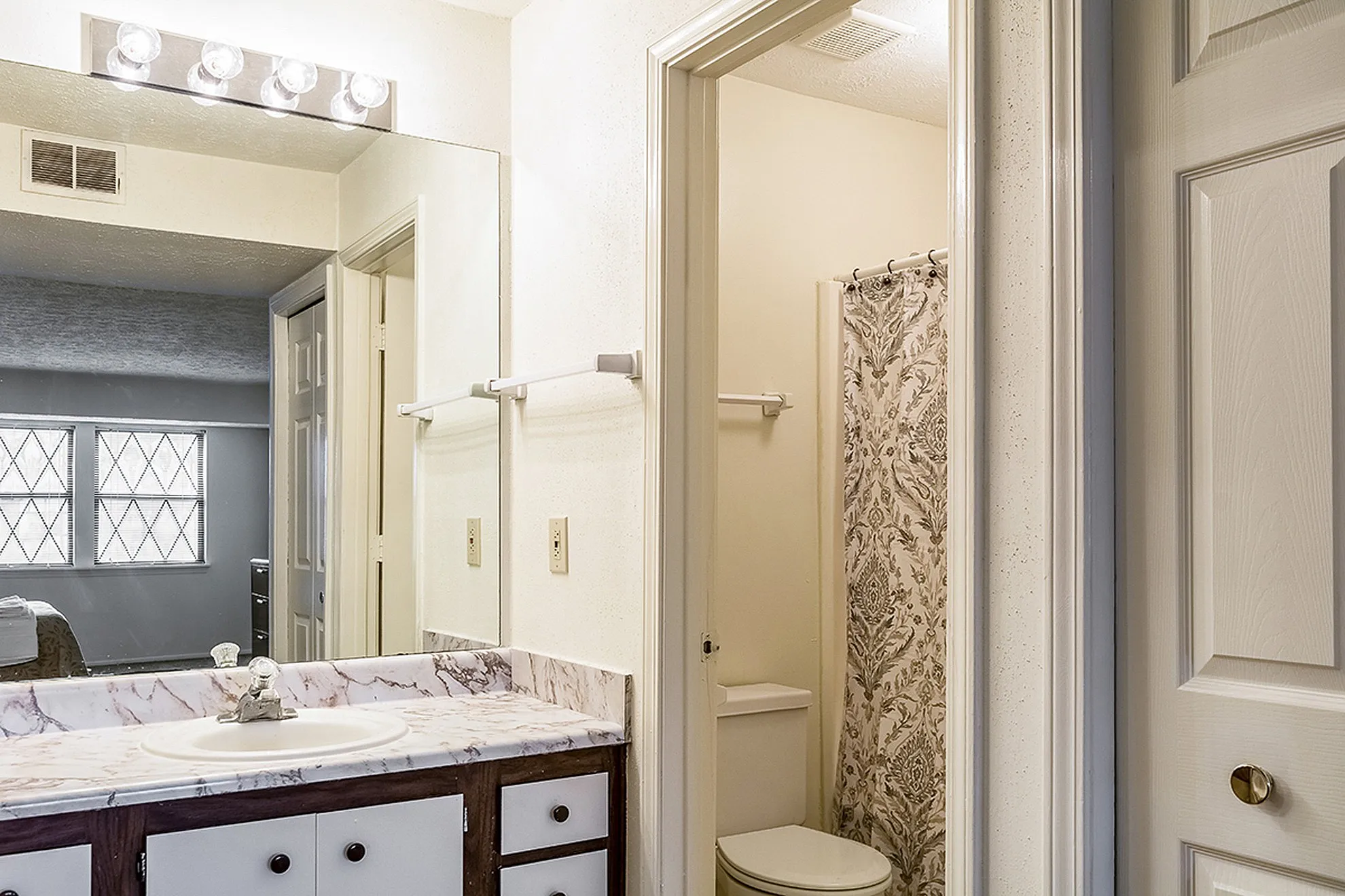 Bright bathroom featuring a large mirror, elegant marble countertop, and floral shower curtain, reflecting modern style.