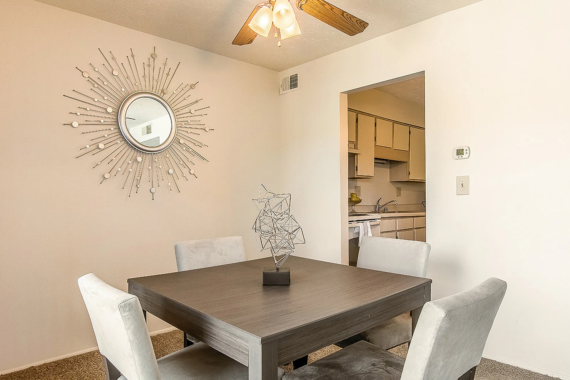 Charming dining area with a unique sunburst wall mirror, modern wooden table, and cozy chairs, leading to a compact kitchen.