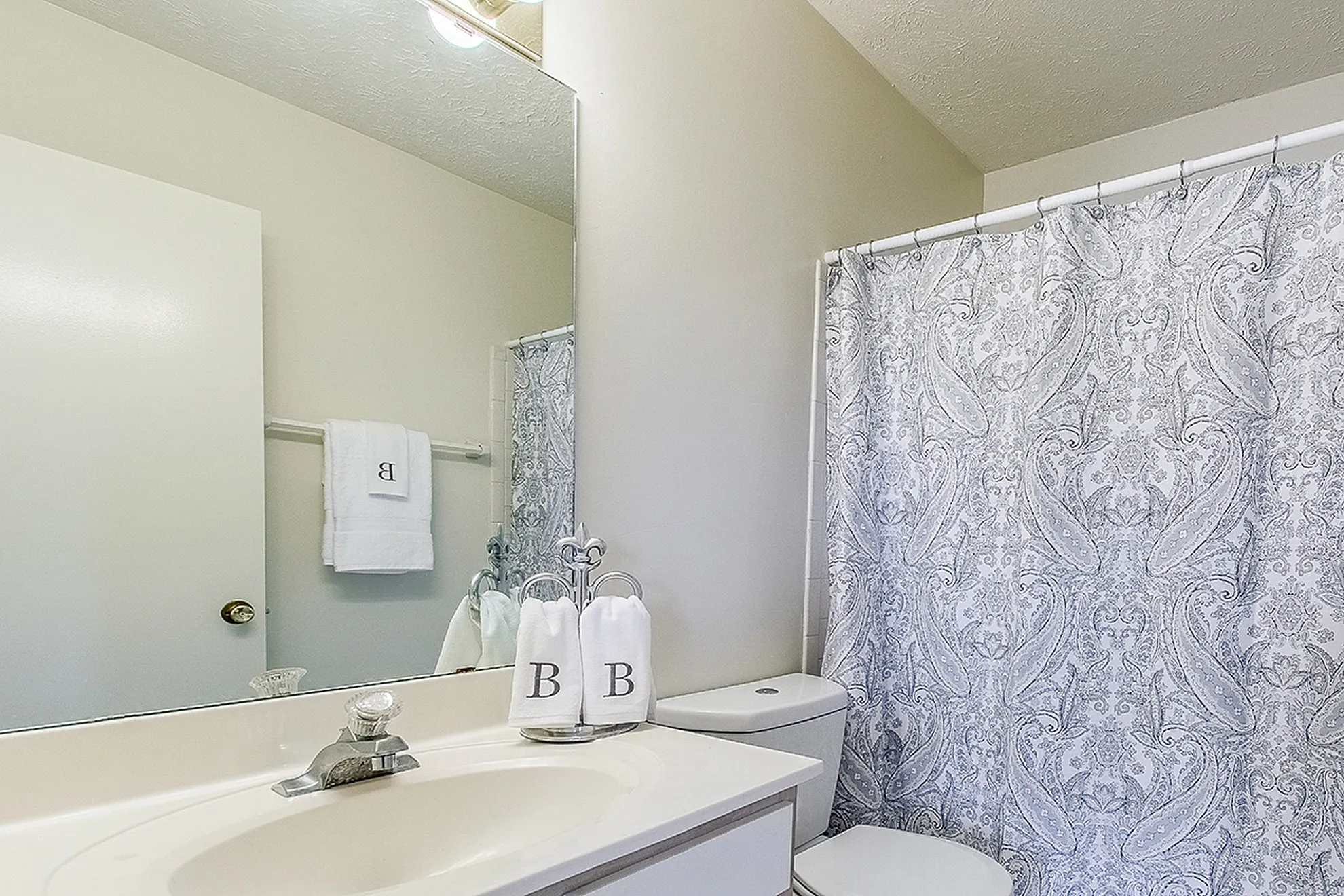 Elegant bathroom featuring a sleek vanity with a large mirror, stylish paisley shower curtain, and coordinated monogrammed towels enhancing a spa-like atmosphere.