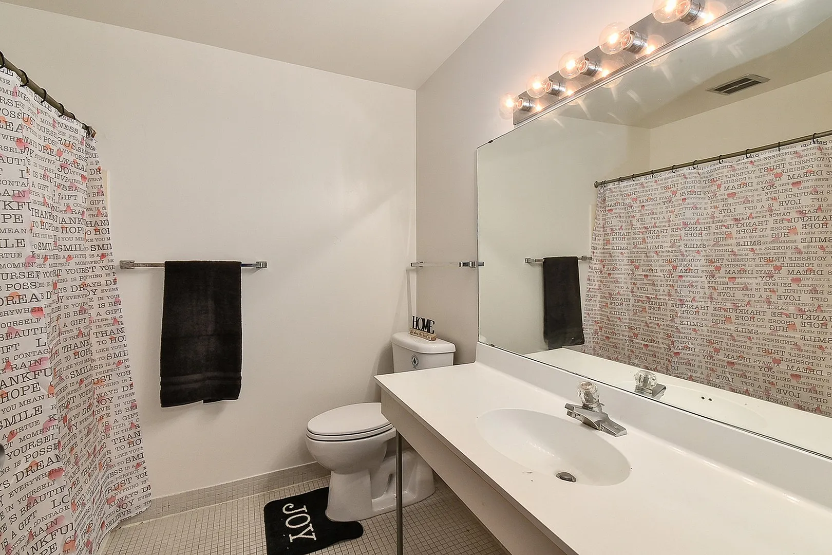 Bright bathroom with a large mirror, modern lighting, and playful shower curtain. Features a sleek white vanity and stylish dark towel accents.