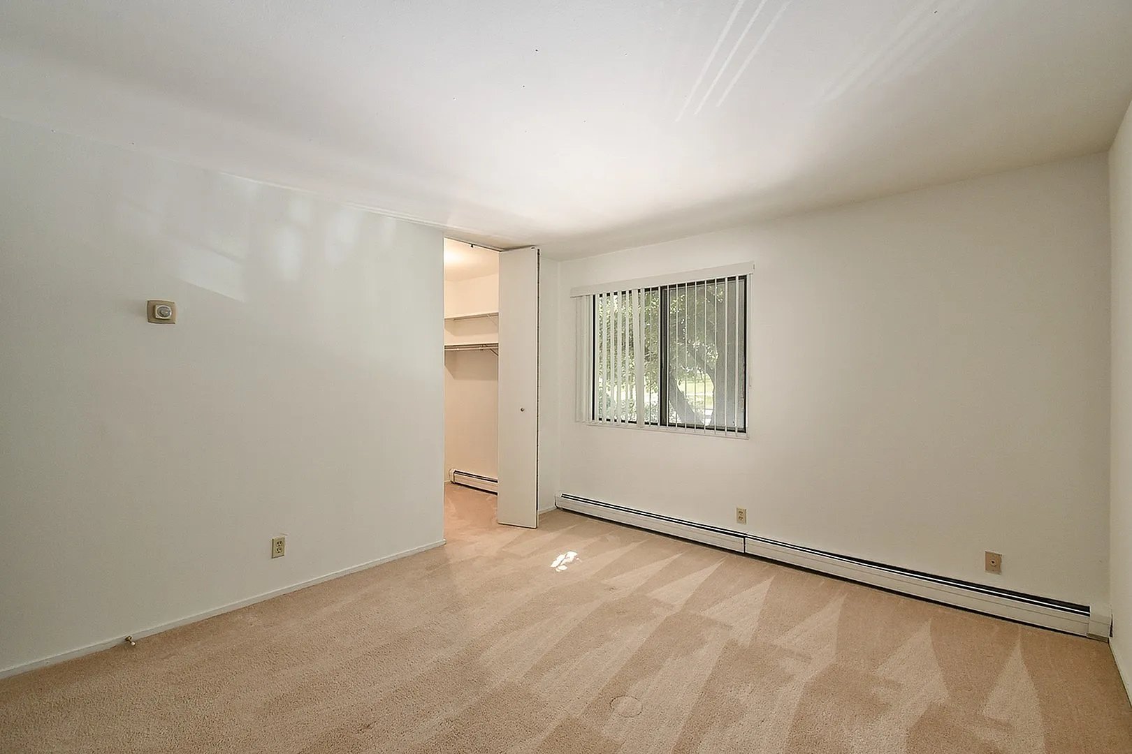Bright bedroom with cozy beige carpet, large window with vertical blinds, and a spacious closet, offering natural light and comfort.