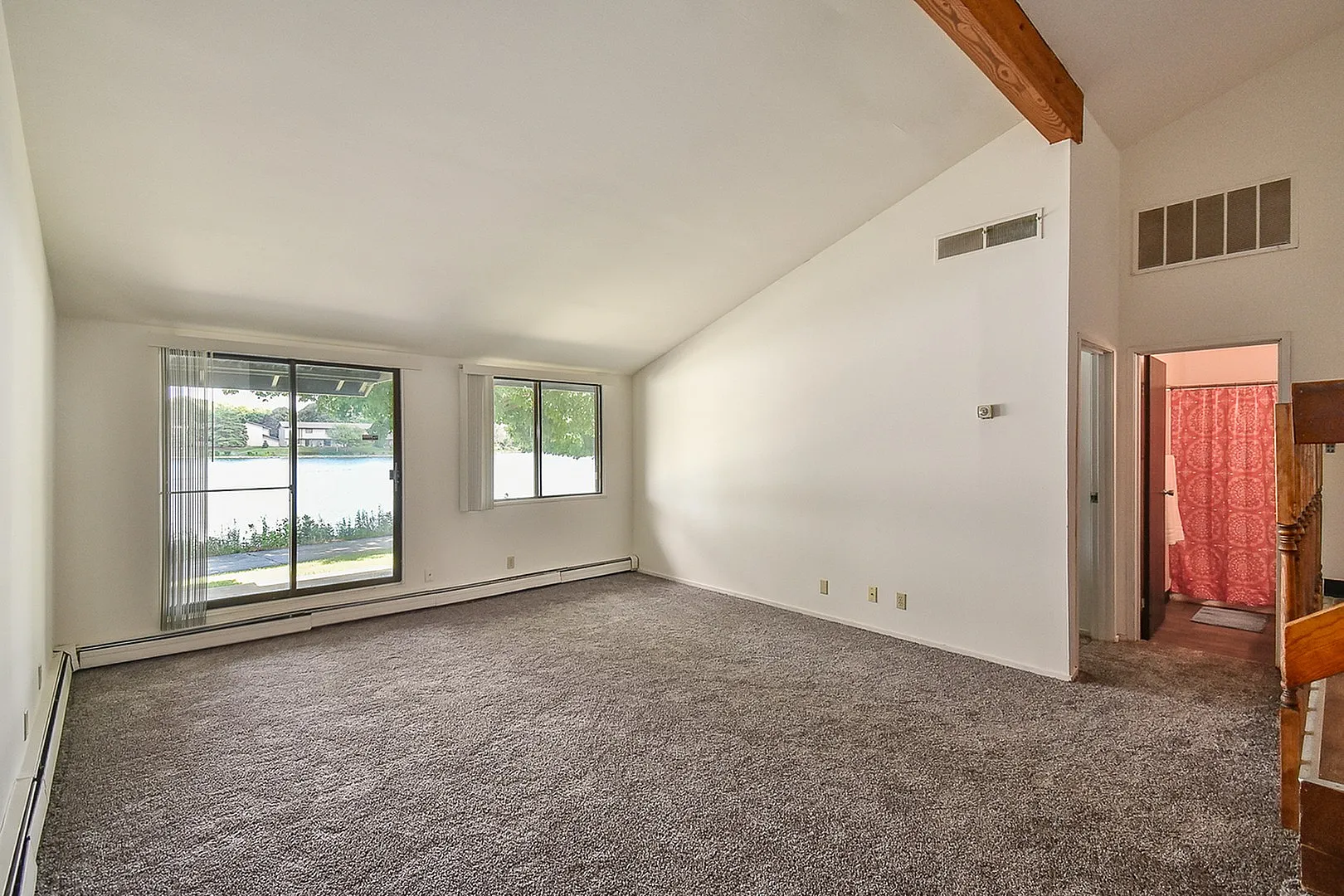 Bright and spacious living room with large windows offering serene waterfront views, featuring plush carpet and open entry to a cozy bathroom area.