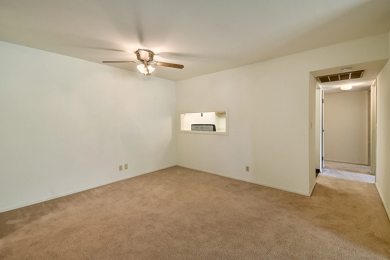 Inviting living area with warm carpet flooring, a ceiling fan, and a pass-through window to the kitchen, perfect for modern living.