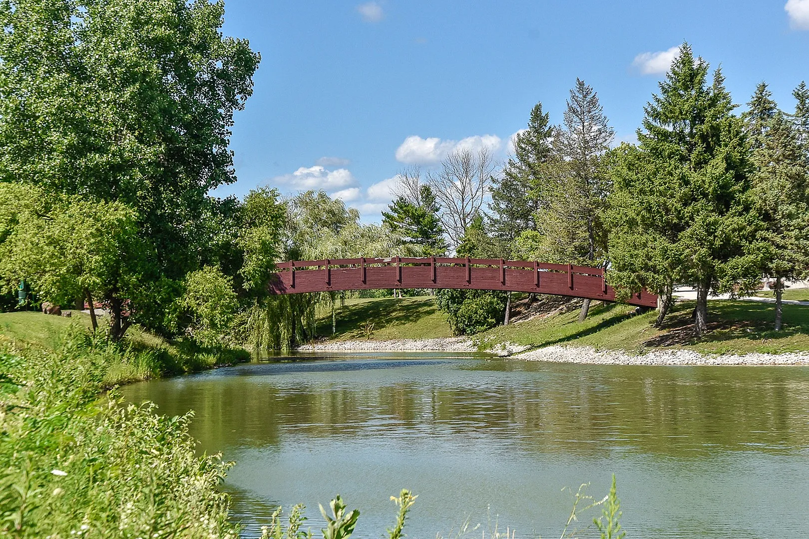 Serene landscape with a charming red bridge arching over a tranquil pond, surrounded by lush green trees under a clear blue sky.