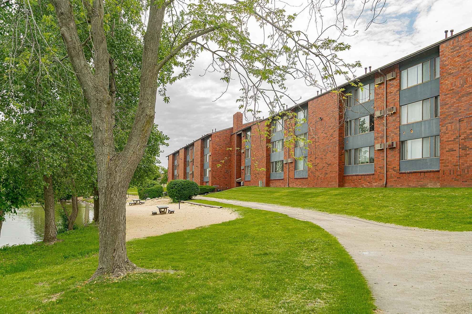 Charming brick apartment building surrounded by lush green landscaping and a scenic walkway, ideal for tranquil living.