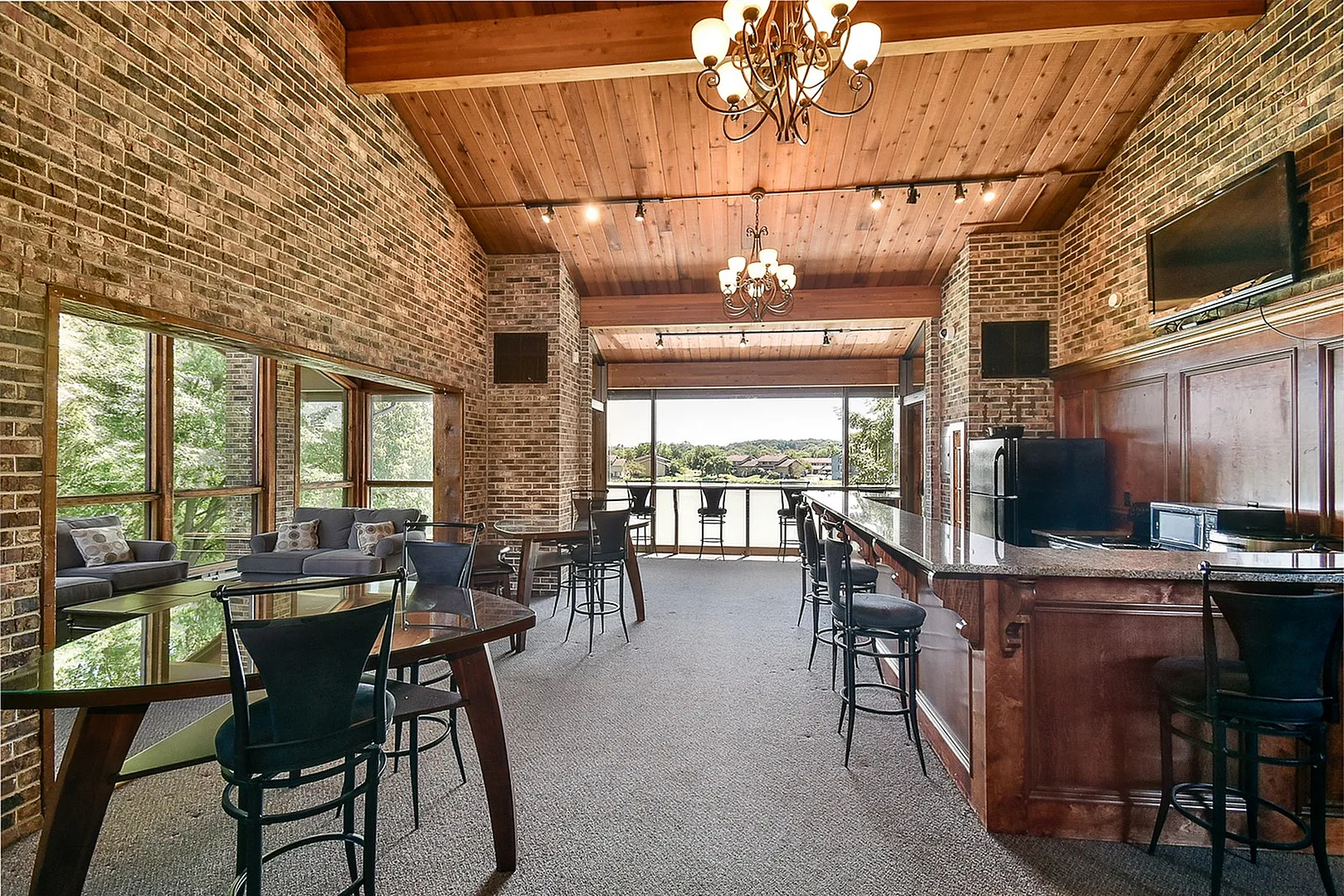 Elegant loft-style lounge with exposed brick walls, large windows overlooking lush greenery, and a spacious bar area.