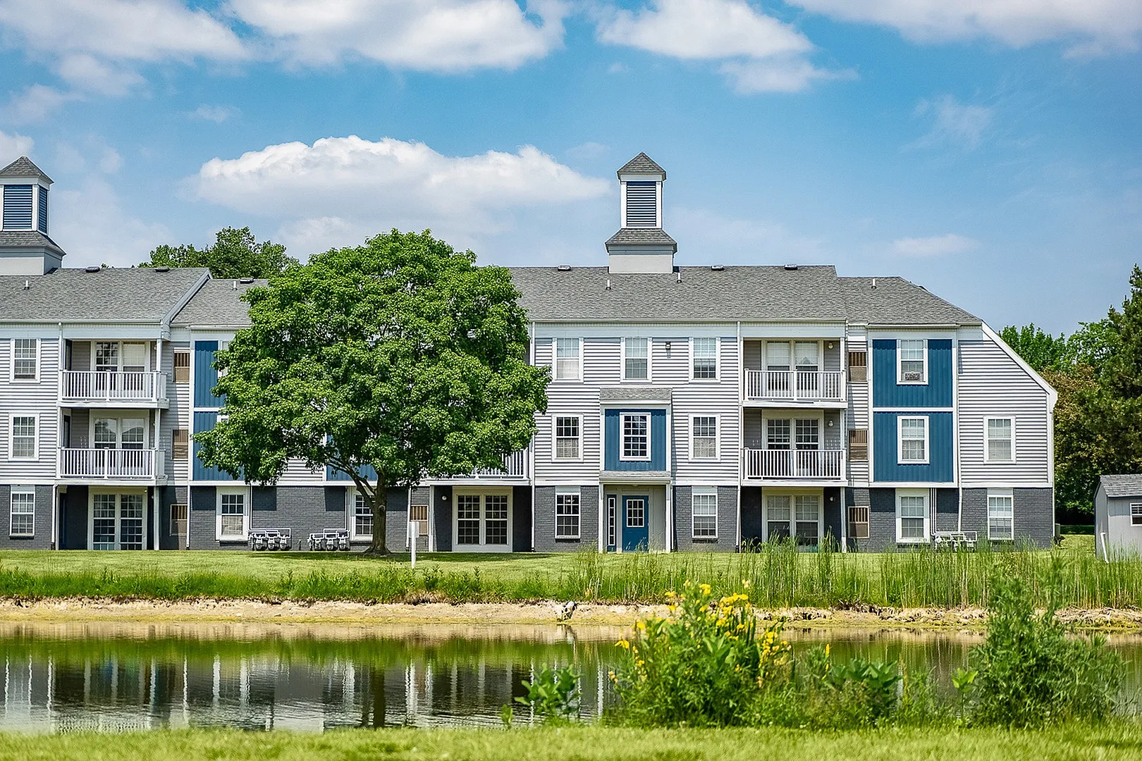 Charming apartment complex with modern architecture, featuring balconies, lush landscaping, and a serene pond view under a blue sky.