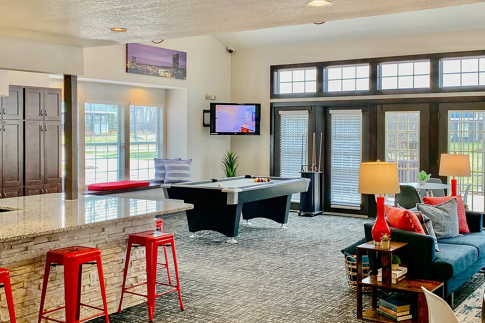 Modern open-concept living area featuring a sleek countertop, vibrant red stools, cozy seating, and a stylish pool table under bright natural light.
