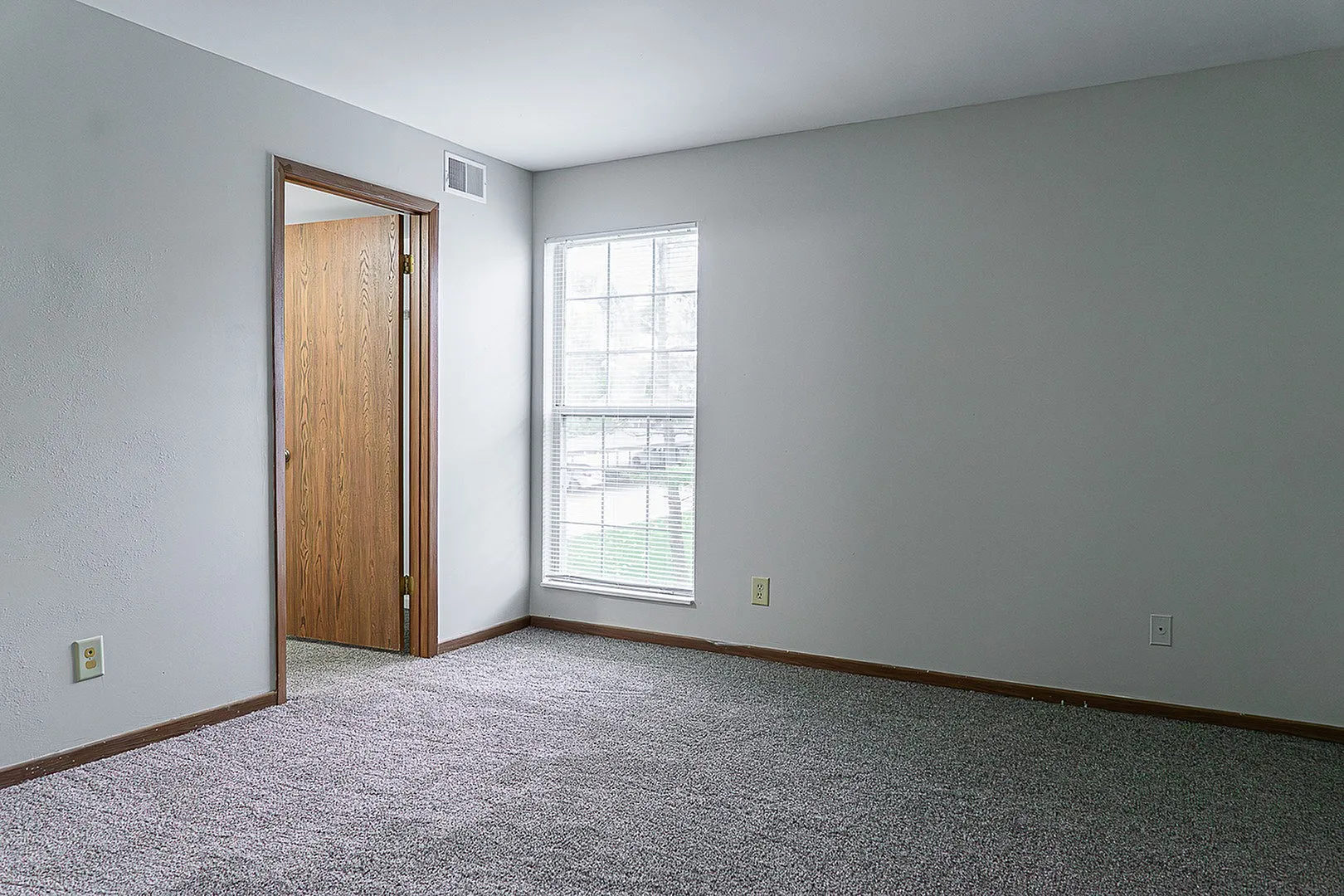Bright and versatile room with soft carpeting, natural light from a large window, and a wooden door, perfect for a cozy bedroom or office space.