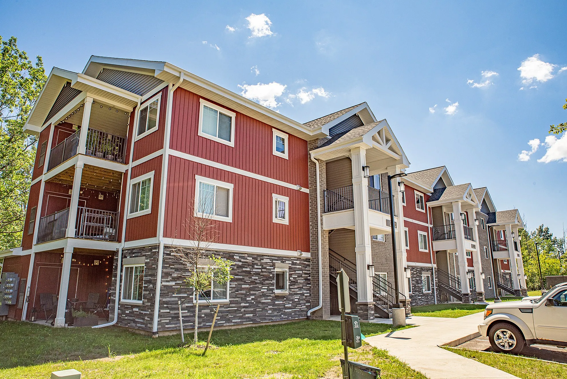 Modern apartment complex featuring stylish red and beige exterior, spacious balconies, and lush green surroundings under a clear blue sky.