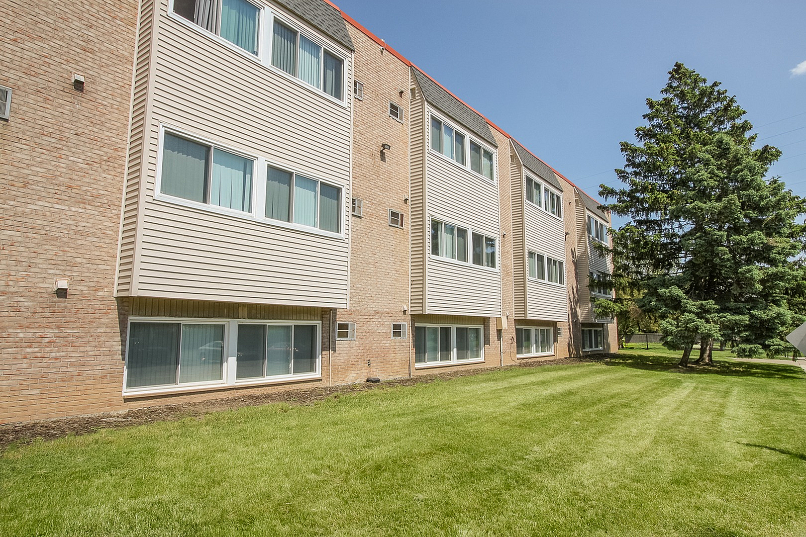 Charming apartment building with a brick facade and large windows, surrounded by lush green grass and a beautiful tree under a clear blue sky.