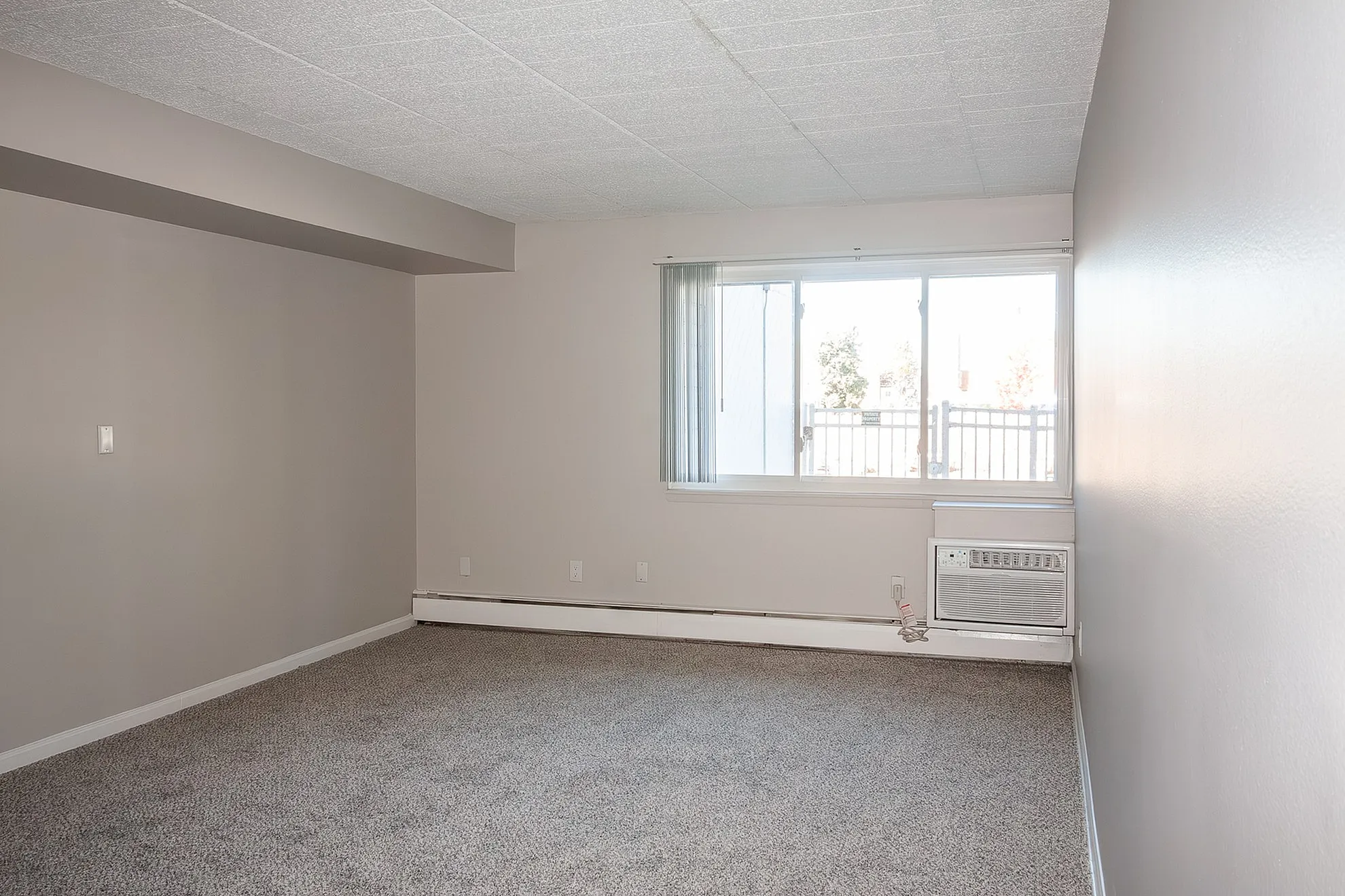 Bright and airy living room with plush carpet, large window, and built-in air conditioner, perfect for a comfortable and inviting space.