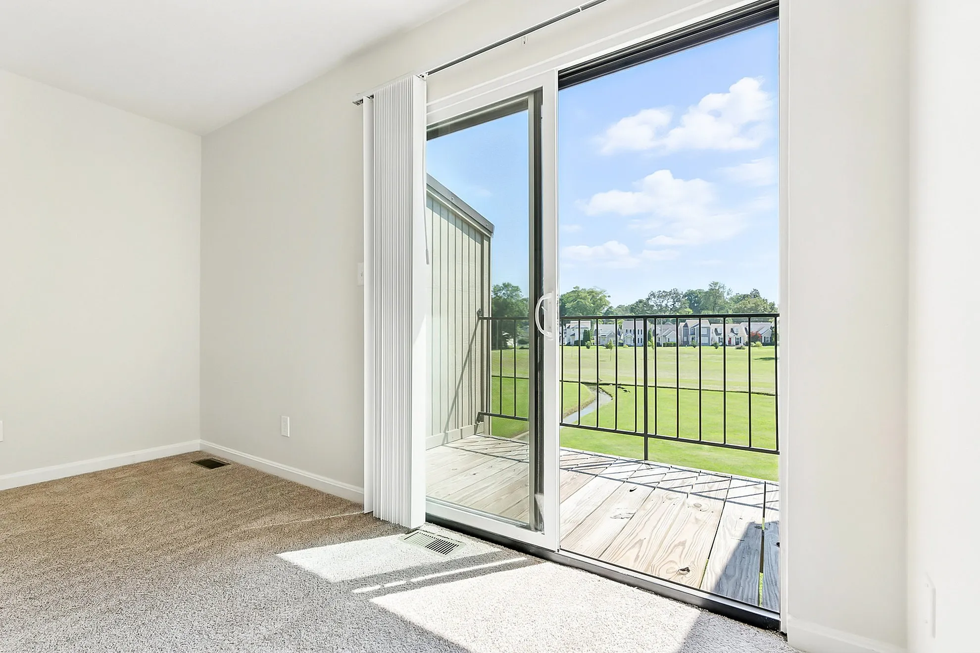 Bright apartment room with sliding glass door leading to a sunny balcony overlooking a lush green landscape.