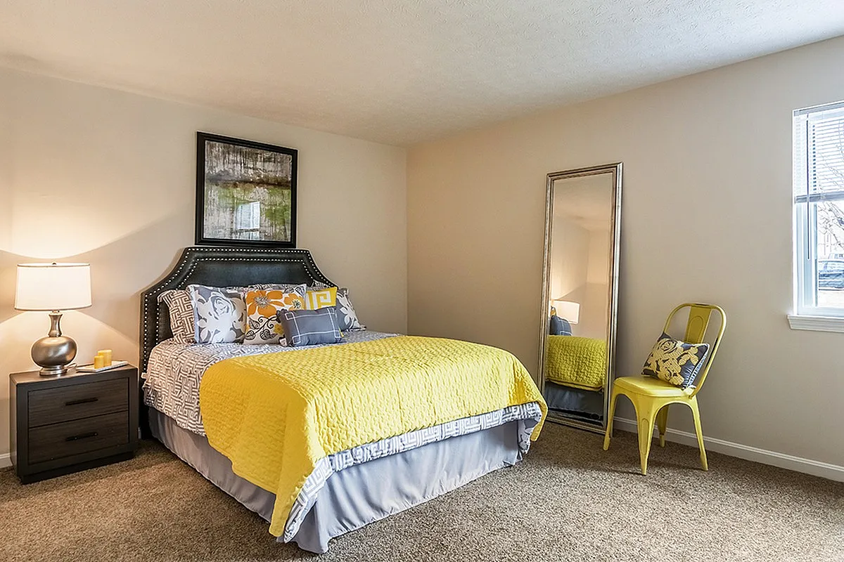 Bright and inviting bedroom with a luxurious bed adorned with vibrant yellow bedding, elegant side lamp, and a stylish full-length mirror beside a decorative chair.