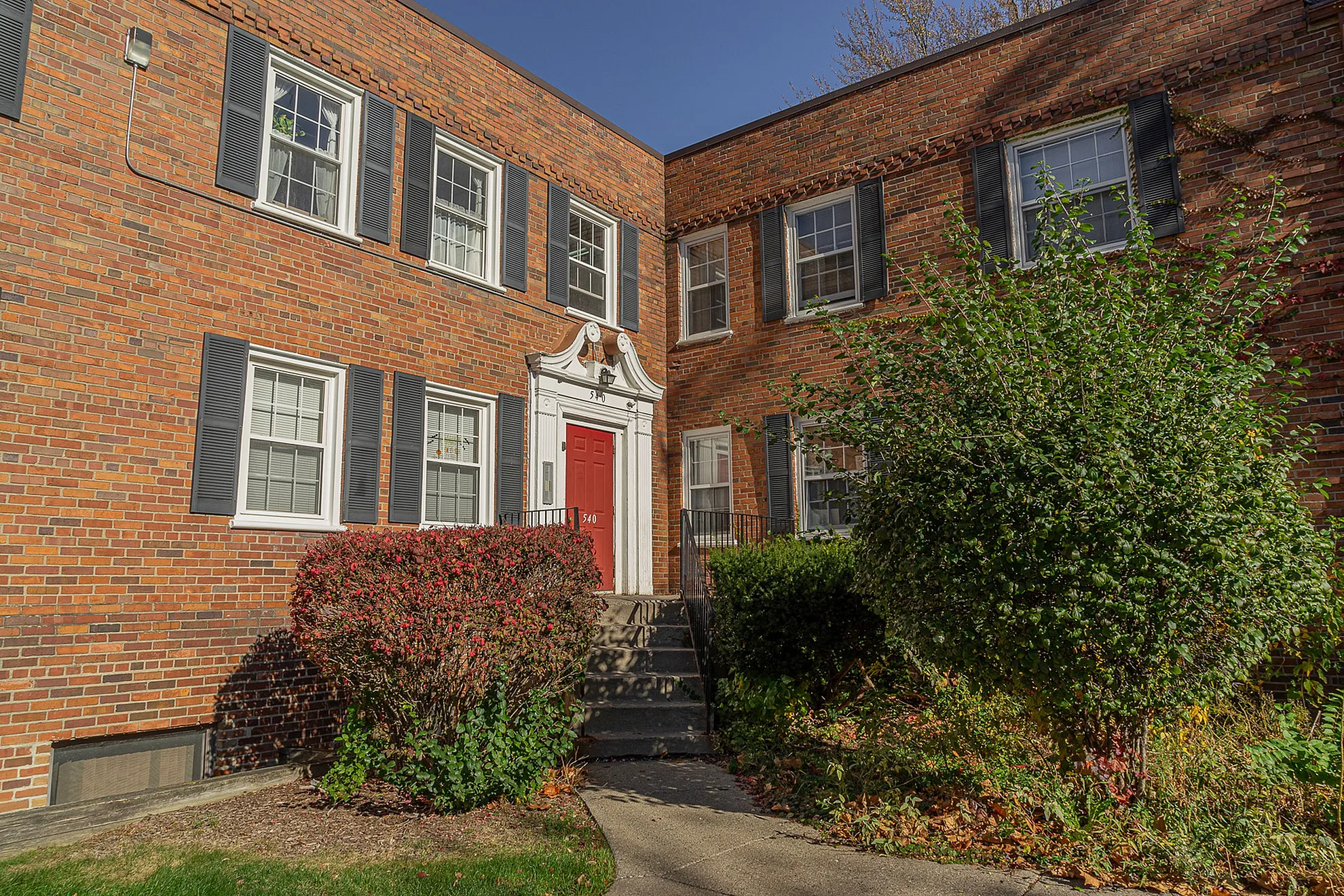 Charming brick apartment building with classic design, featuring a vibrant red door, stylish shutters, and lush greenery.