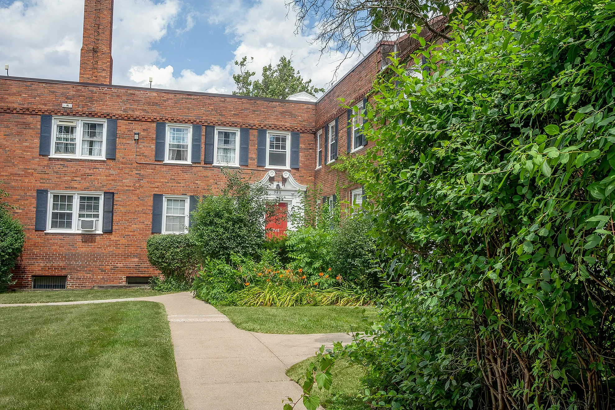 Charming brick apartment complex with vibrant green landscaping and a welcoming entrance under a bright blue sky.