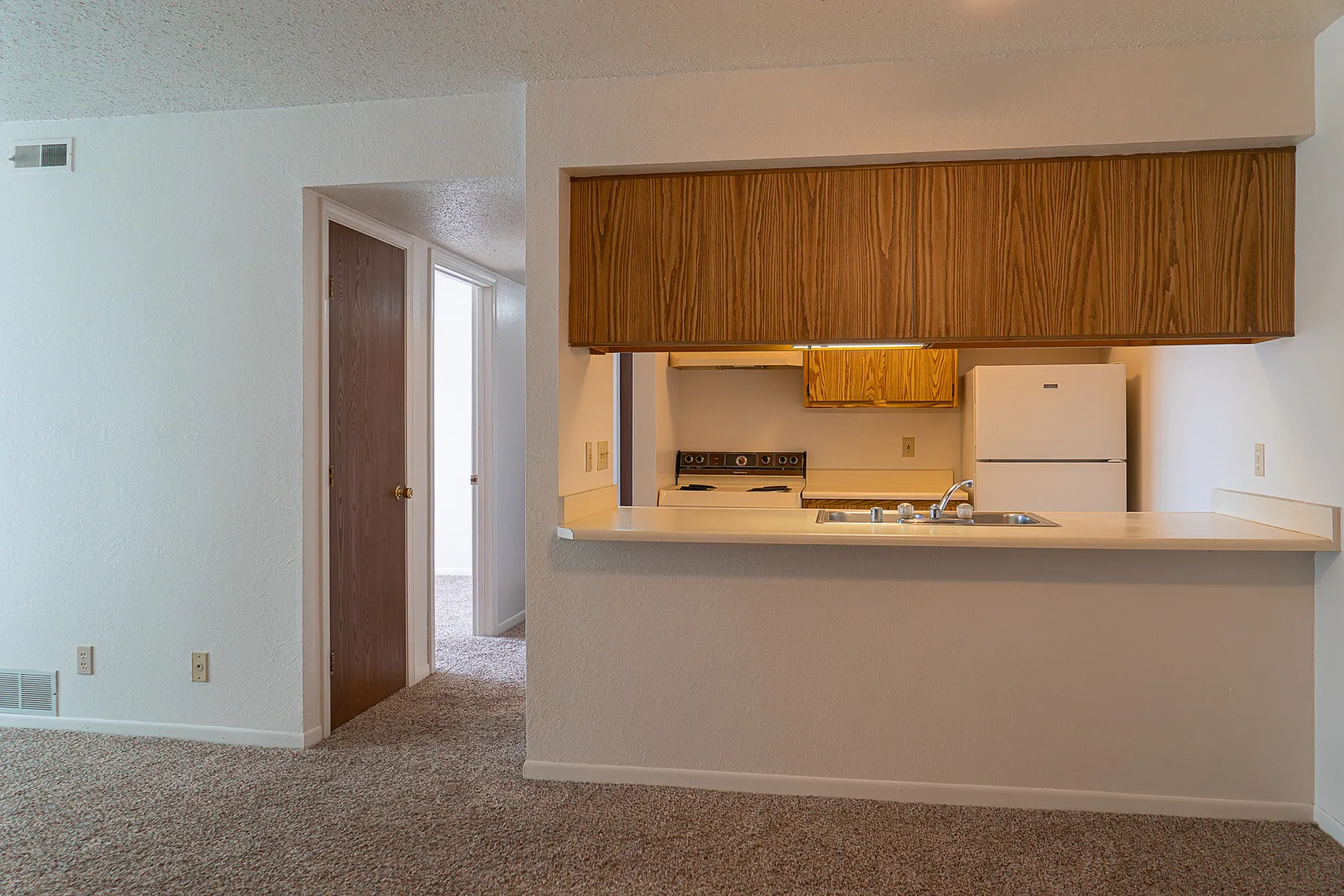 Inviting open kitchen layout with warm wooden cabinets and modern appliances. Cozy entrance leads to carpeted spaces.
