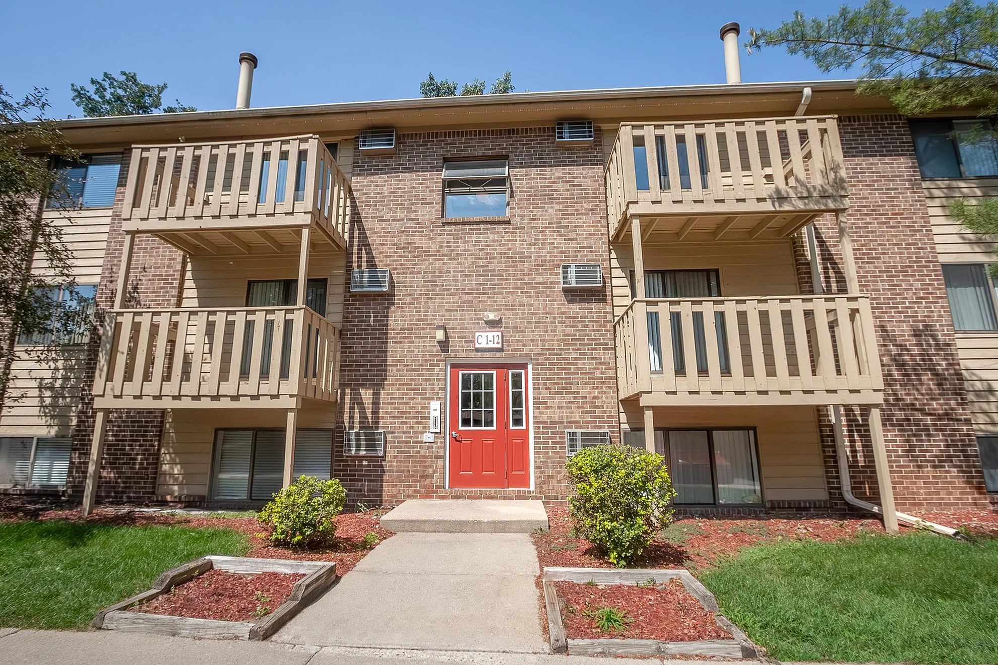 Charming apartment building with a brick exterior, inviting balconies, and a bright red door, set amidst leafy surroundings.