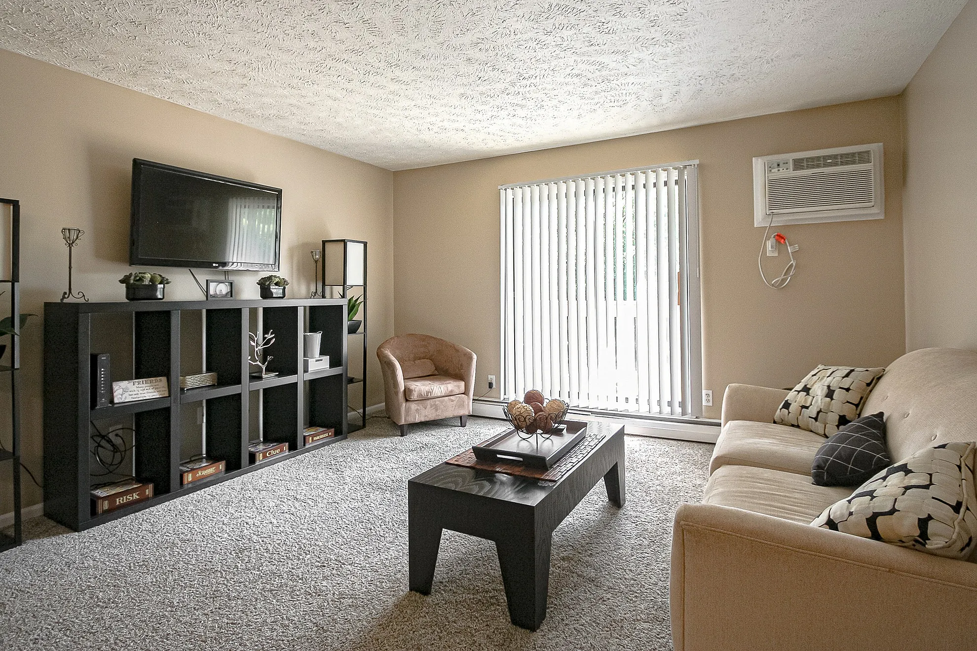 Bright and inviting living room with plush carpeting, sleek black entertainment center, cozy seating, and abundant natural light from large vertical blinds.