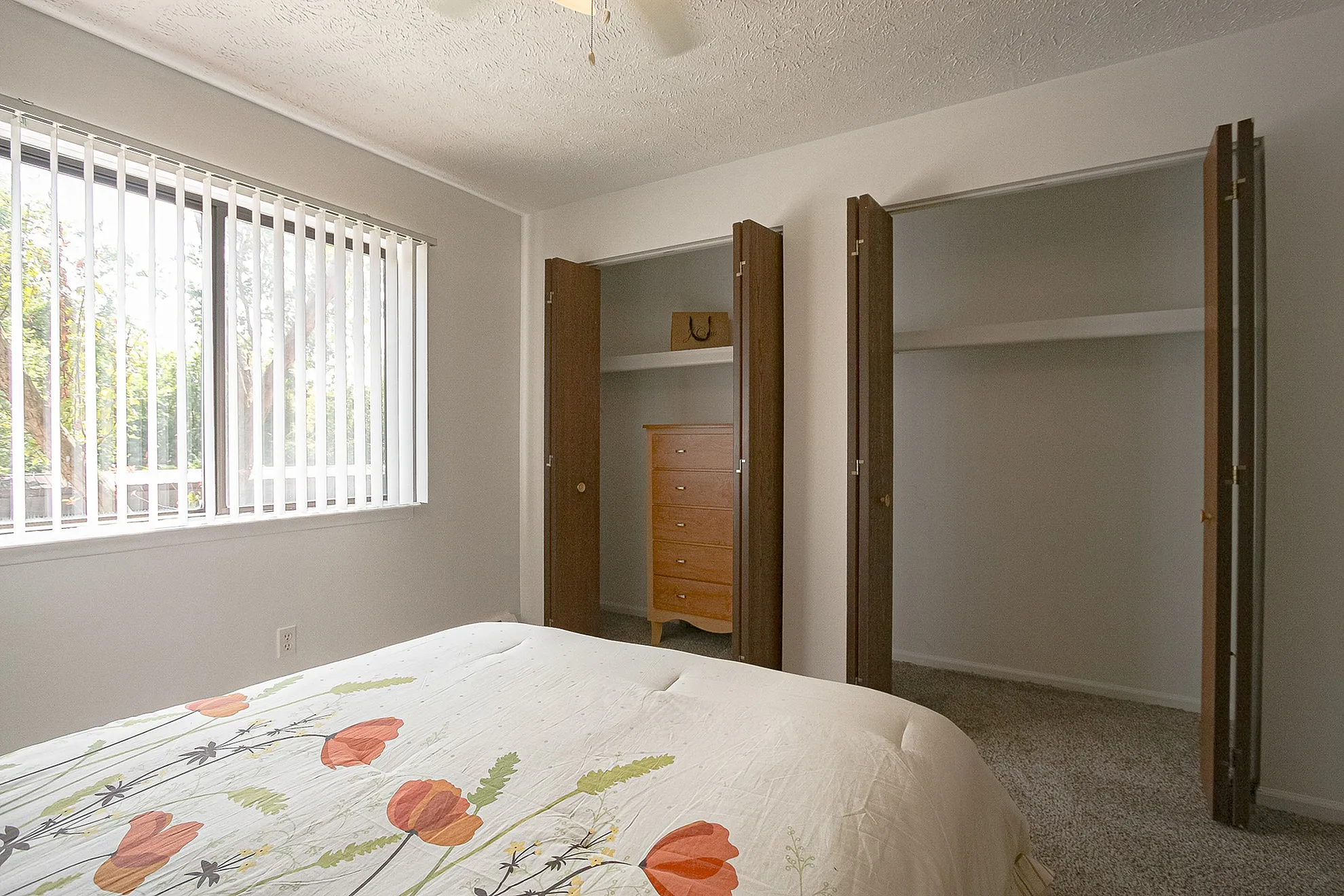 Bright bedroom with floral bedding, large windows, dual closets, and soft carpet flooring.