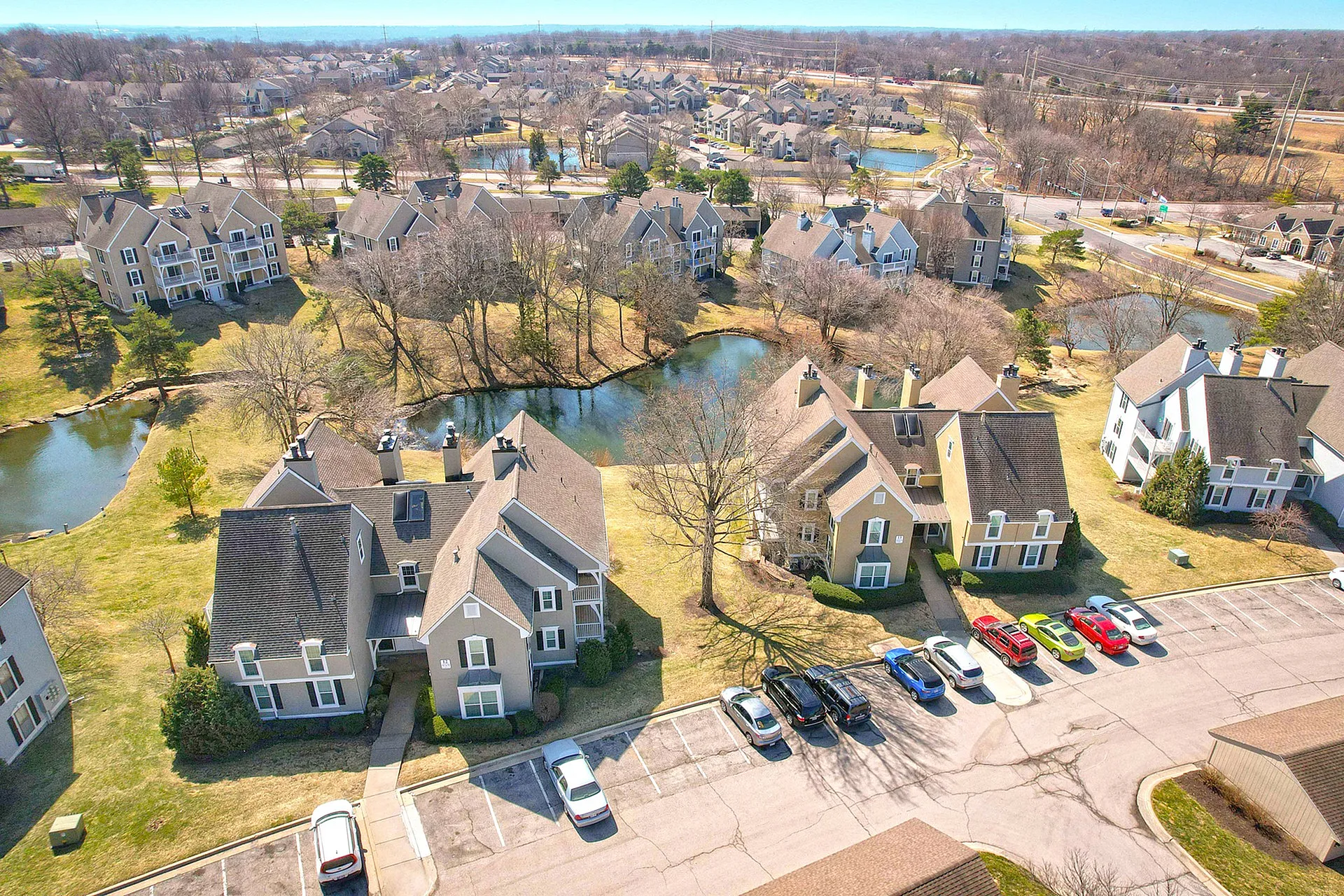 Scenic aerial view of charming apartment complex surrounded by lush greenery and serene ponds, featuring spacious parking and tranquil residential environment.