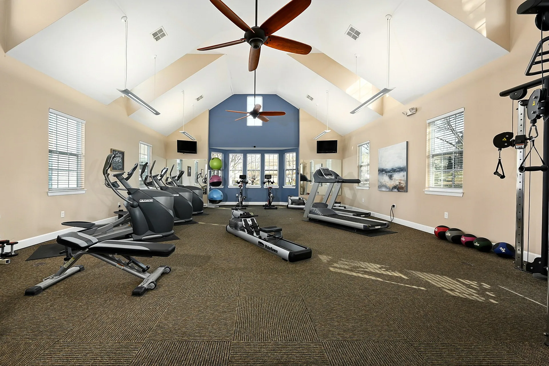 Bright, spacious fitness center featuring modern exercise equipment, including treadmills, rower, elliptical machines, and free weights, under high ceilings with ceiling fans.