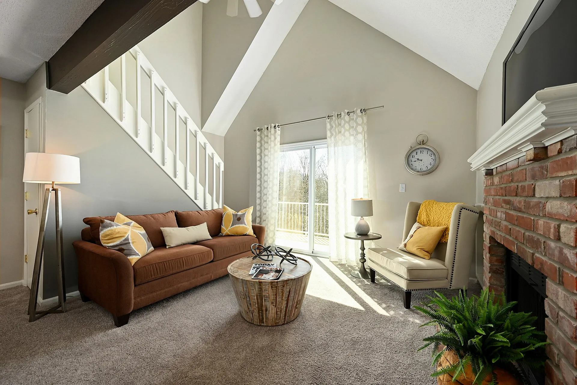 Bright and cozy living room with vaulted ceiling, brick fireplace, and large windows offering natural light, featuring a comfortable brown sofa and elegant armchair.