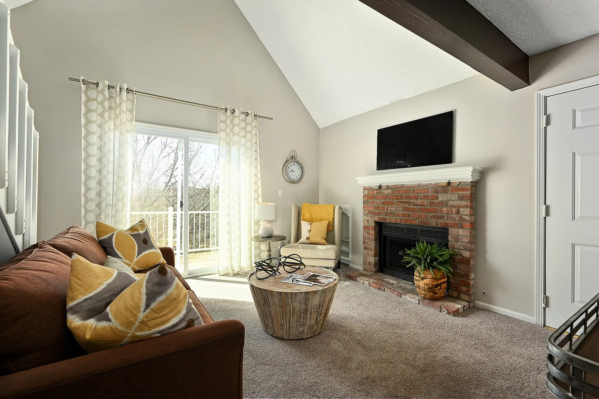 Bright living room with vaulted ceiling, featuring a cozy brick fireplace, plush seating, and large sliding glass doors opening to a scenic balcony.