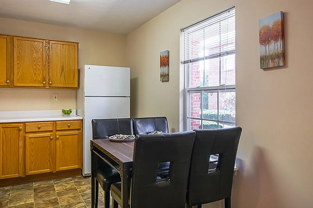 Bright dining area in modern kitchen with elegant wooden cabinetry, sleek white refrigerator, and cozy seating by a sunlit window.
