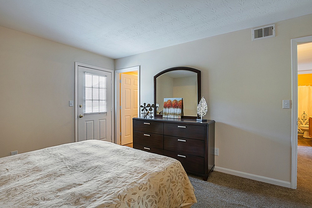 Inviting bedroom featuring a cozy bed, elegant dresser with mirror, and a door leading to a bright, airy space.