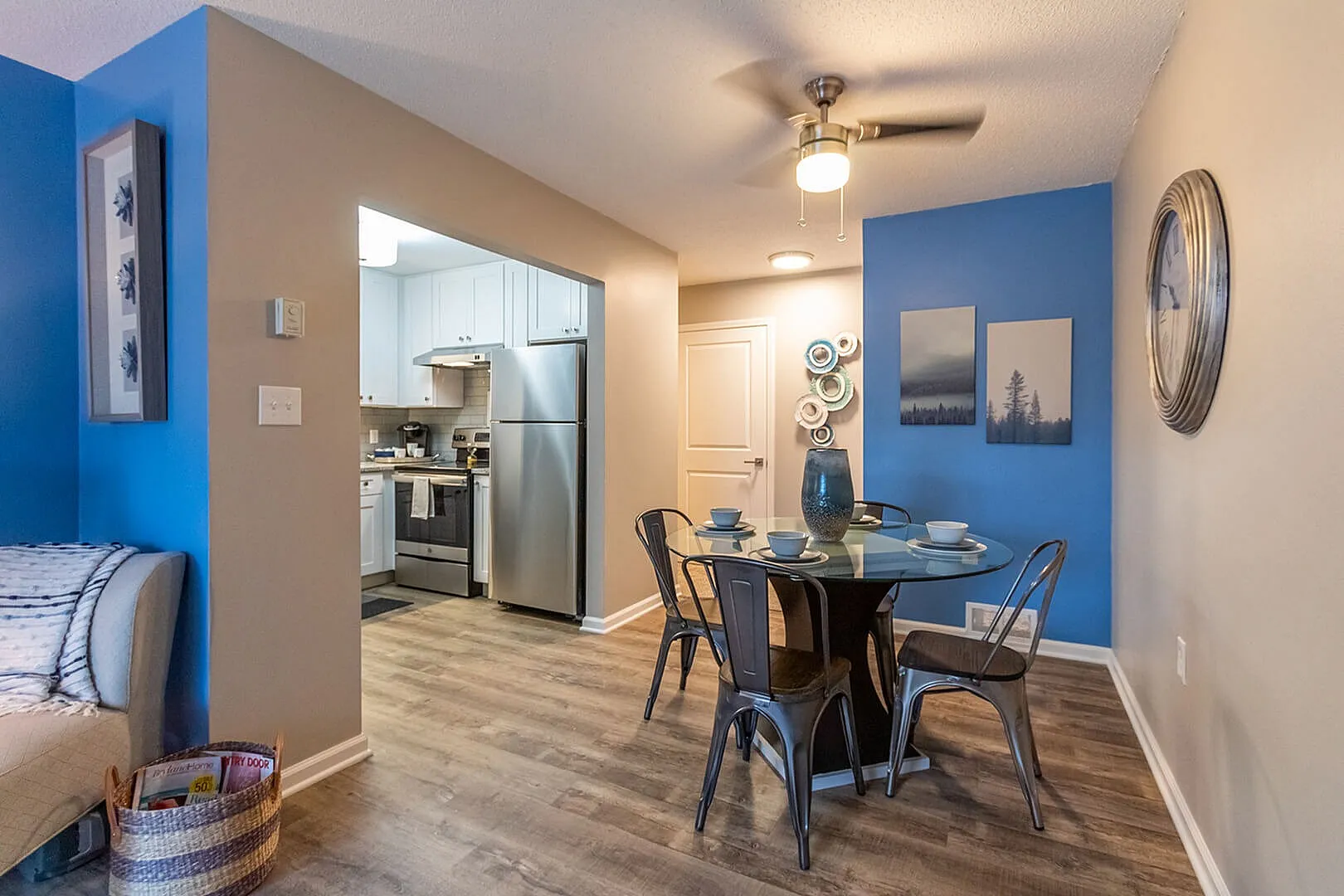 Chic dining area with a glass table set for four, adjoining a modern kitchen with stainless steel appliances, accented by stylish decor and a vibrant blue feature wall.