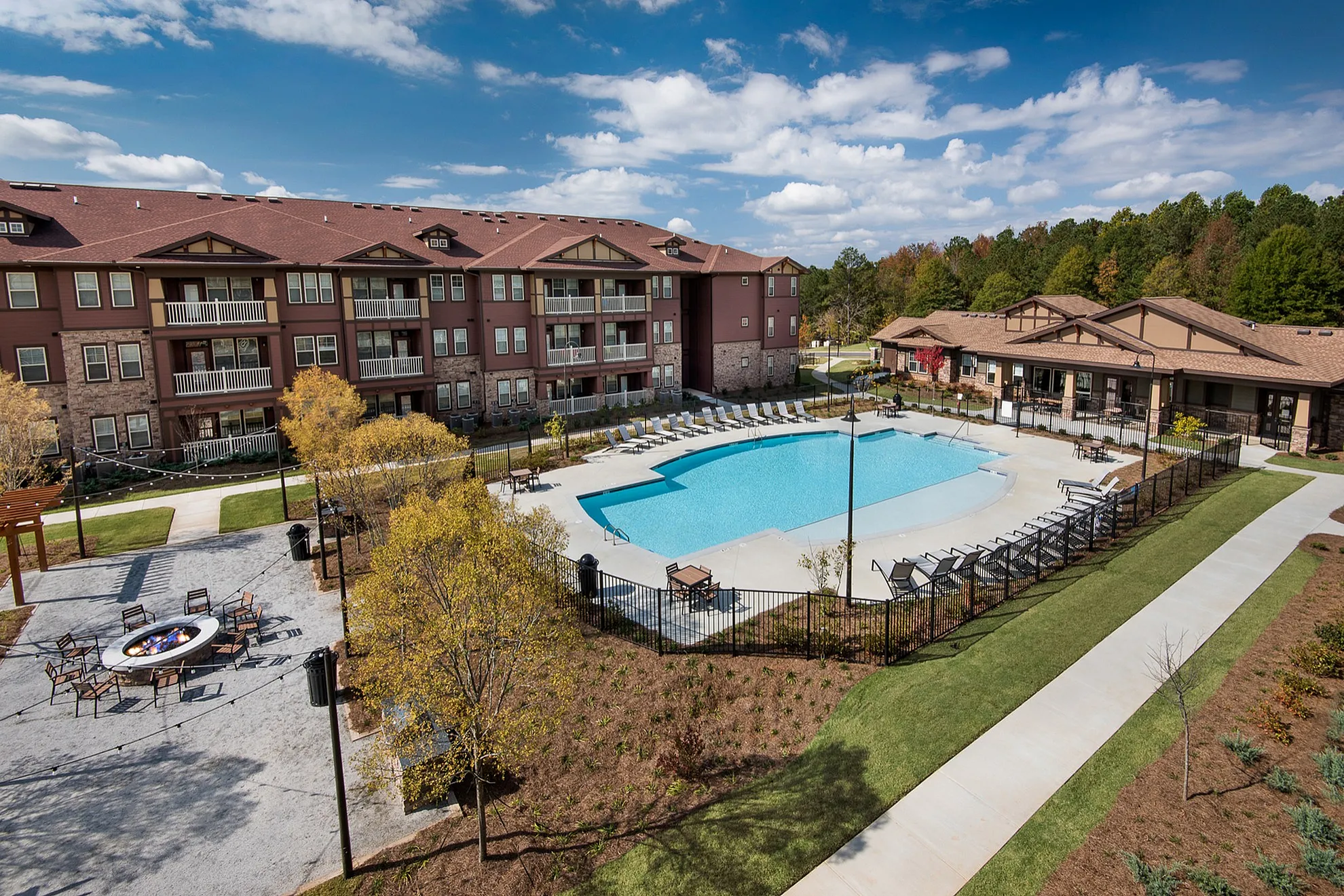 Beautiful apartment complex featuring a sparkling blue pool, outdoor seating area, and lush green landscaping under a bright blue sky, offering a perfect blend of comfort and leisure.