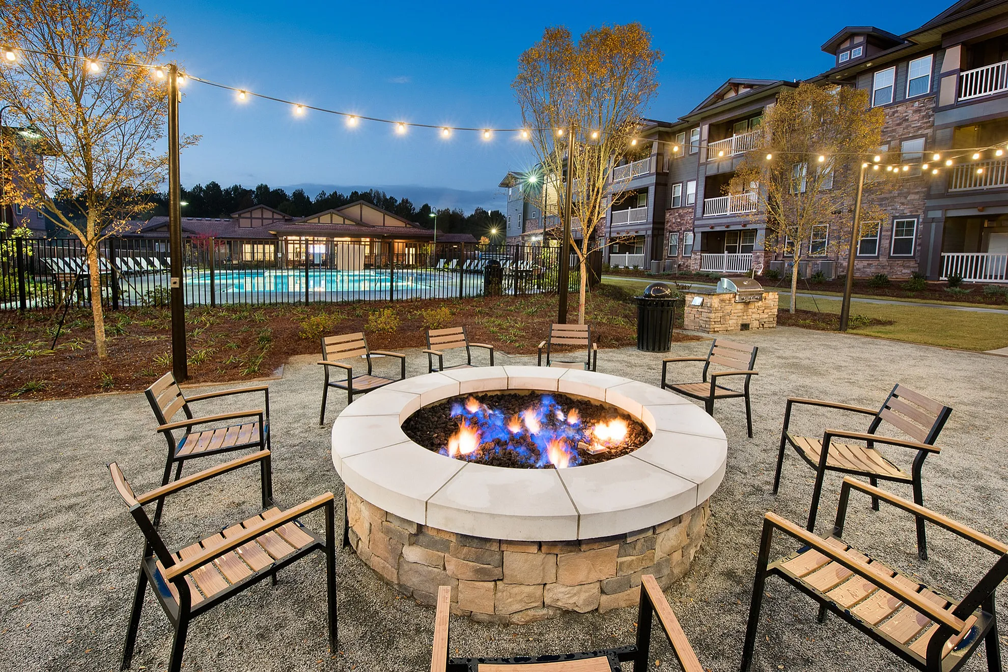 Inviting outdoor space with a warm fire pit, surrounded by modern seating and fairy lights, adjacent to a sparkling pool and stylish apartment complex.