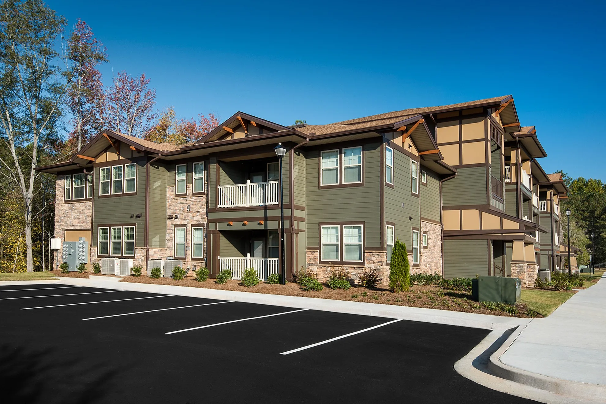 Charming two-story apartment building with modern green siding, stone accents, and inviting balconies, surrounded by lush greenery and ample parking space.