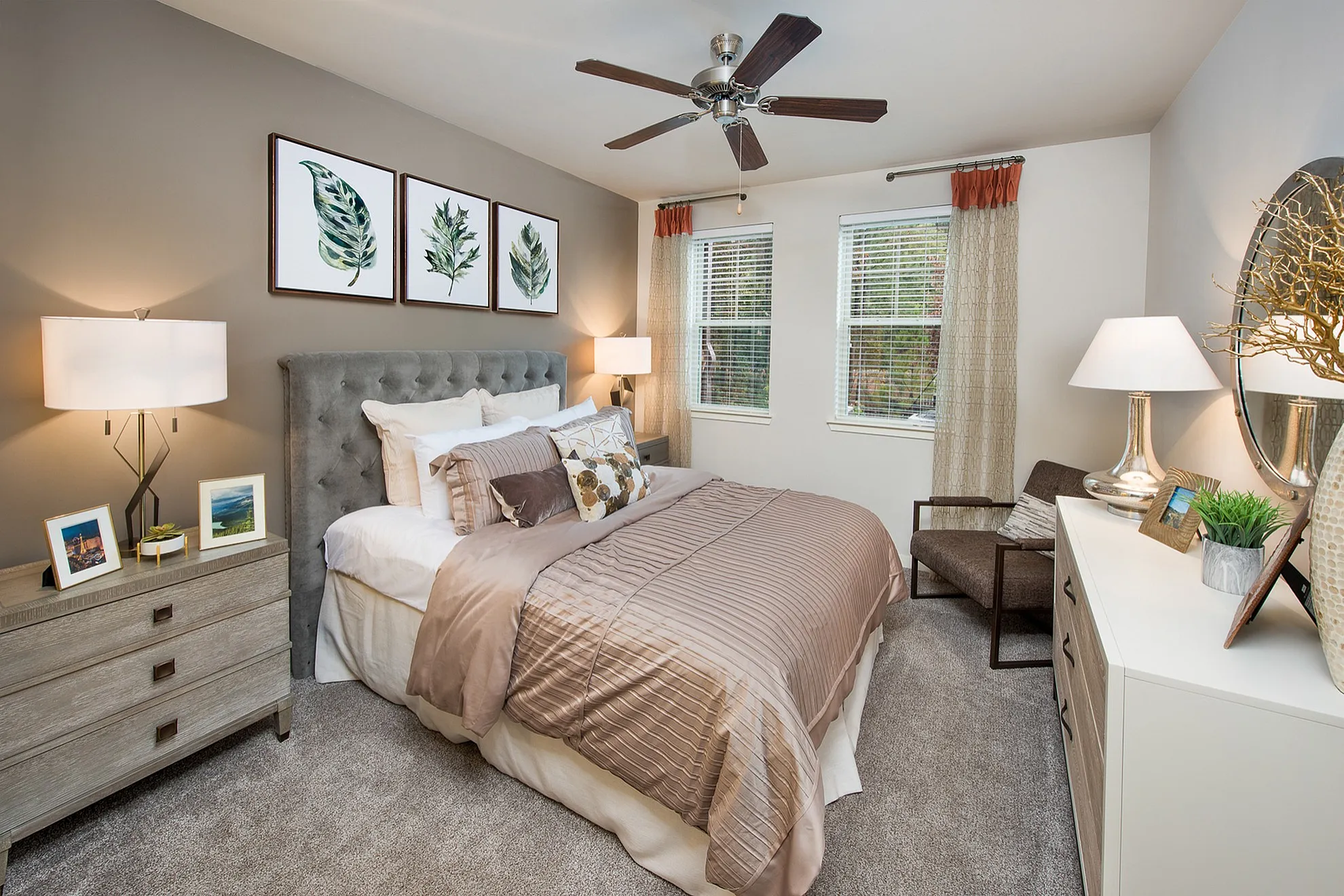 Elegant bedroom with a stylish gray tufted headboard, cozy bedding, and botanical wall art. Features modern lighting, natural light from large windows, and warm accents.