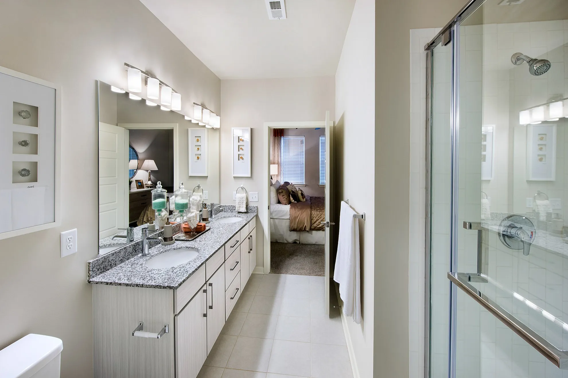 Elegant bathroom with dual sinks, granite countertops, and a modern glass shower, leading into a cozy bedroom.