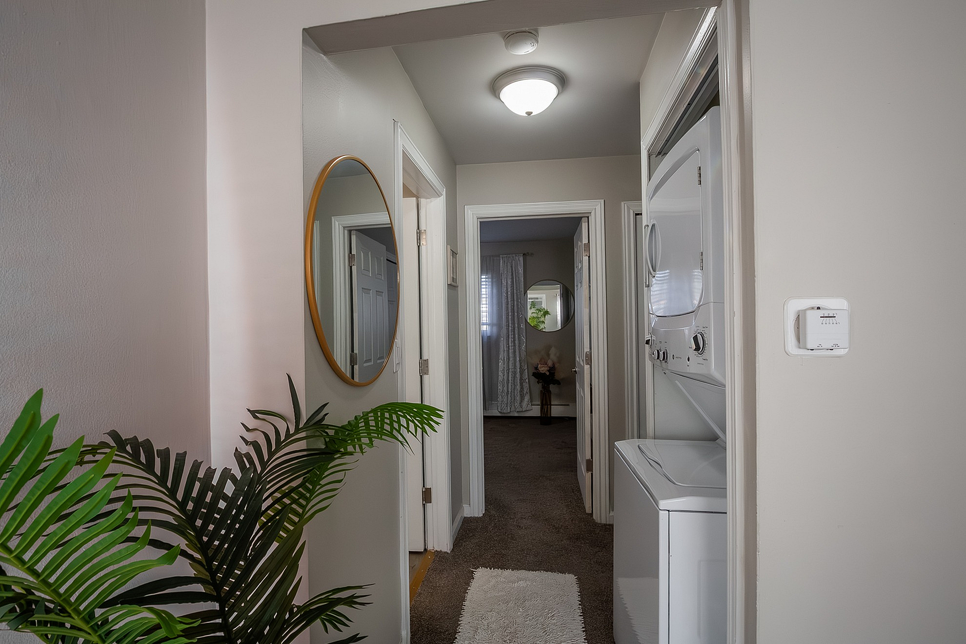 Charming hallway with elegant lighting, a large round mirror, and convenient in-unit laundry, leading to a cozy bedroom with greenery accents.