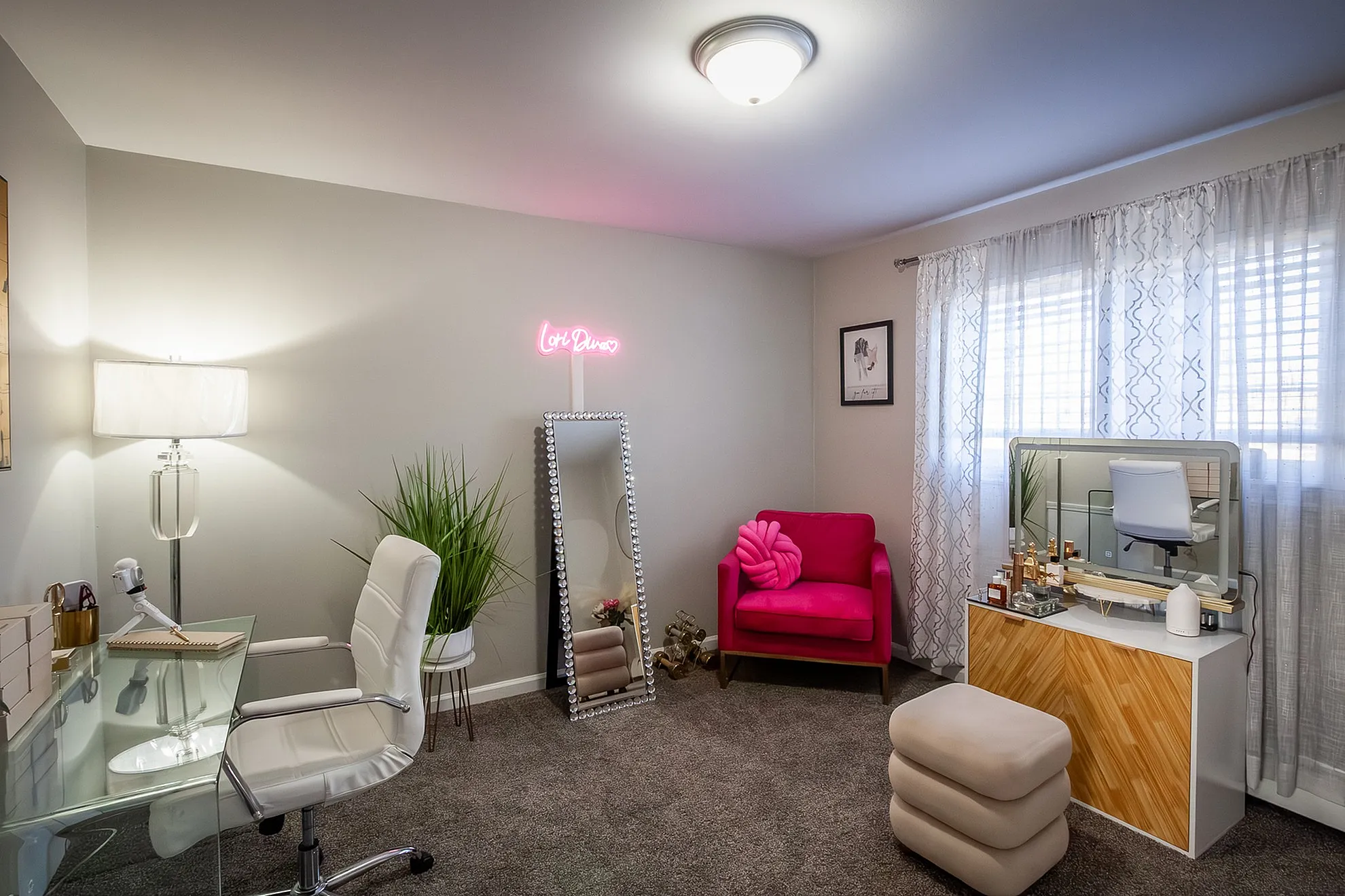 Chic home office with modern glass desk, vibrant pink chair, and elegant decor featuring a neon sign and full-length mirror.