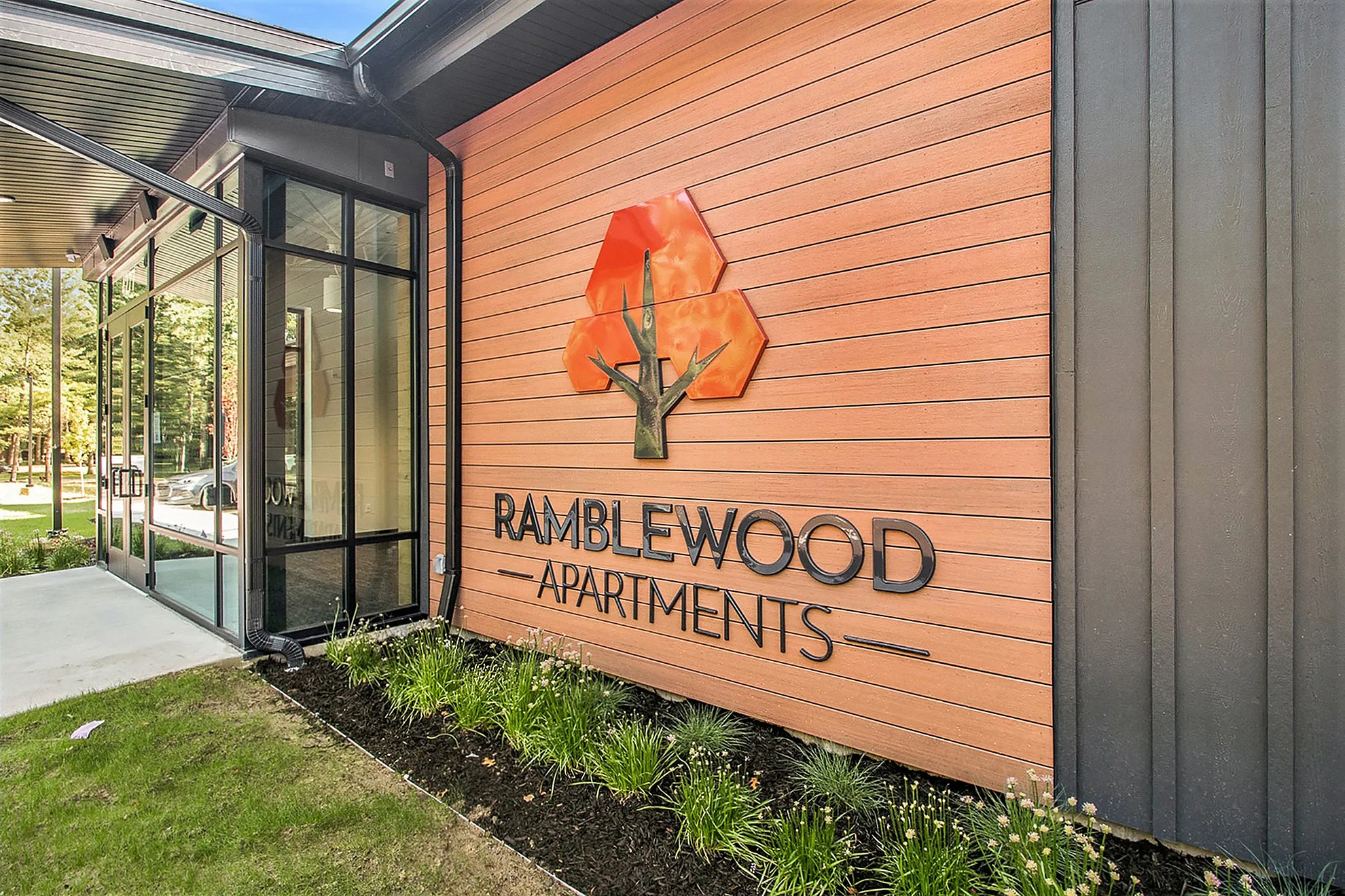 Modern entrance to Ramblewood Apartments with stylish wooden exterior and unique logo, surrounded by lush greenery.