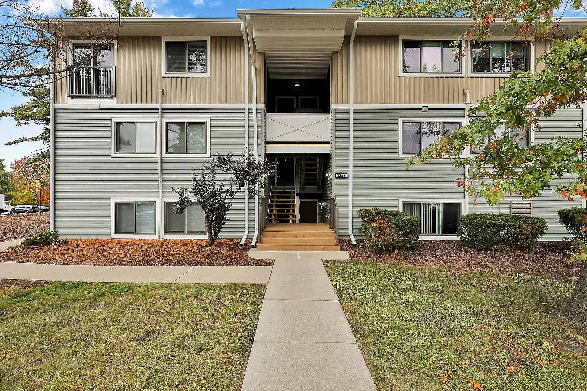 Charming apartment building with modern siding, surrounded by lush greenery and vibrant trees, offering a welcoming entrance.