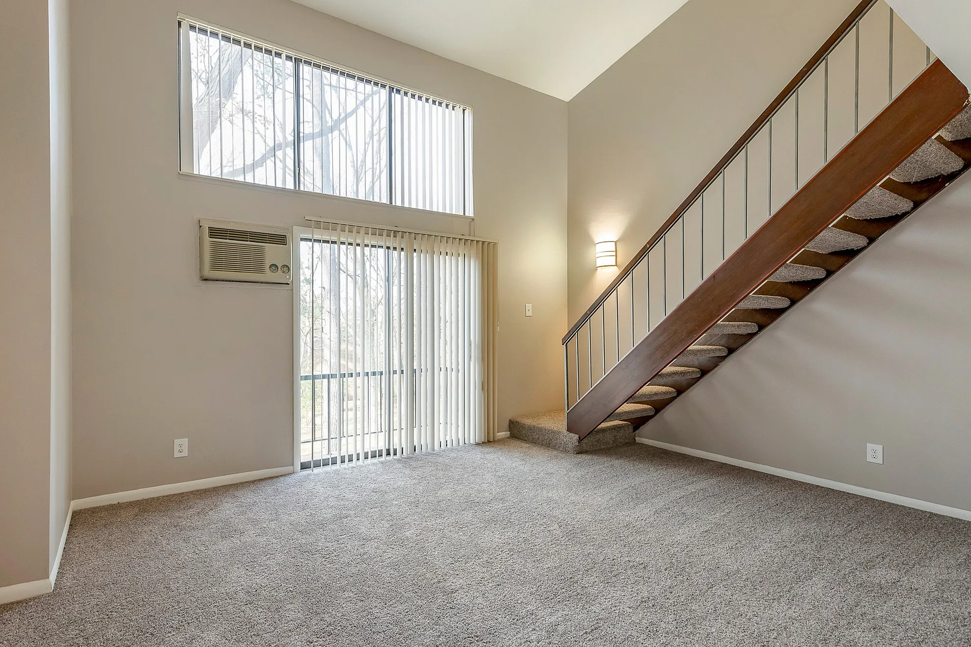 Bright and inviting living space with plush carpet, high ceilings, large windows, and a sleek wooden staircase.