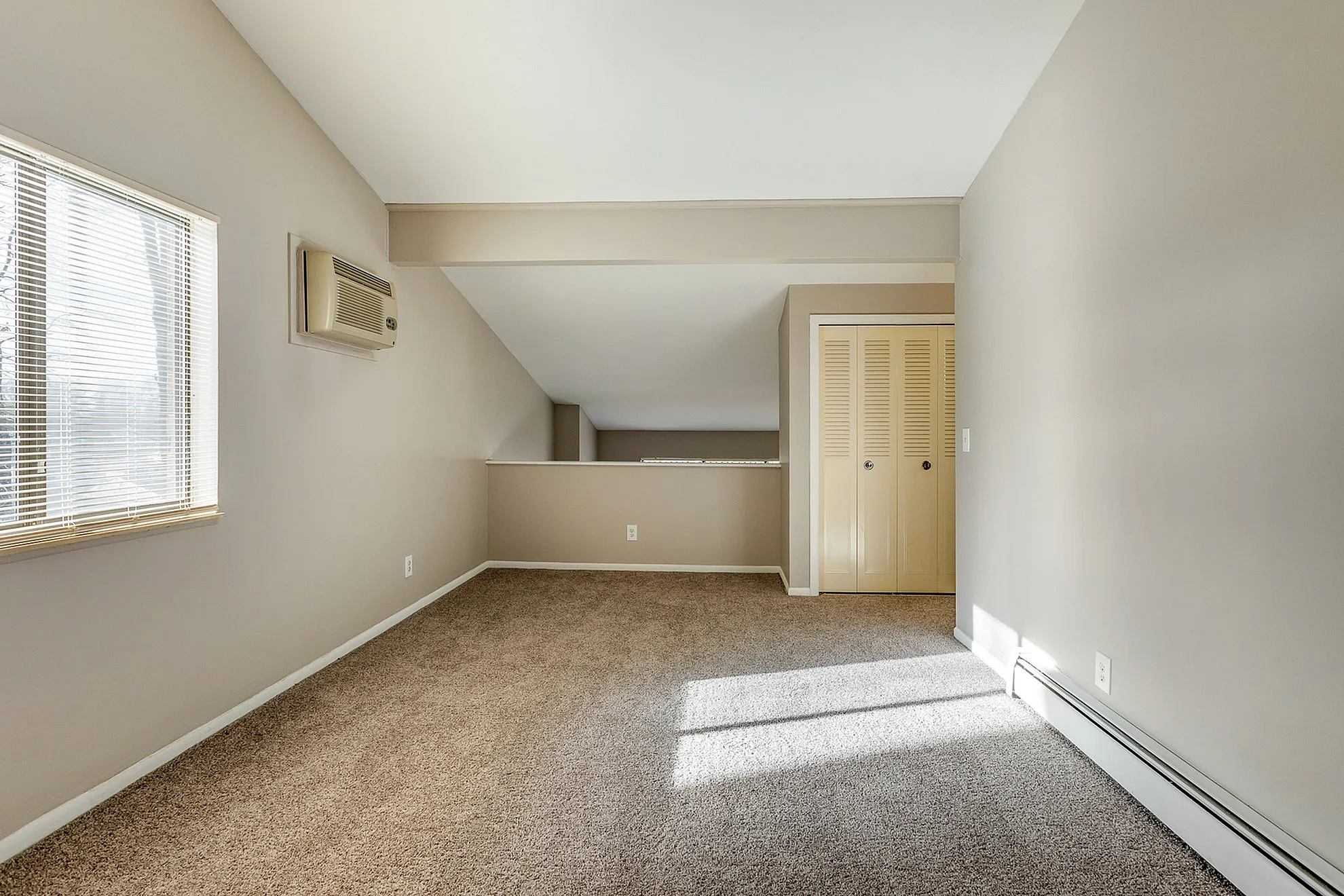 Bright and cozy room with plush carpet flooring, ample natural light from a window, and a sleek air conditioning unit, perfect for relaxing at home.