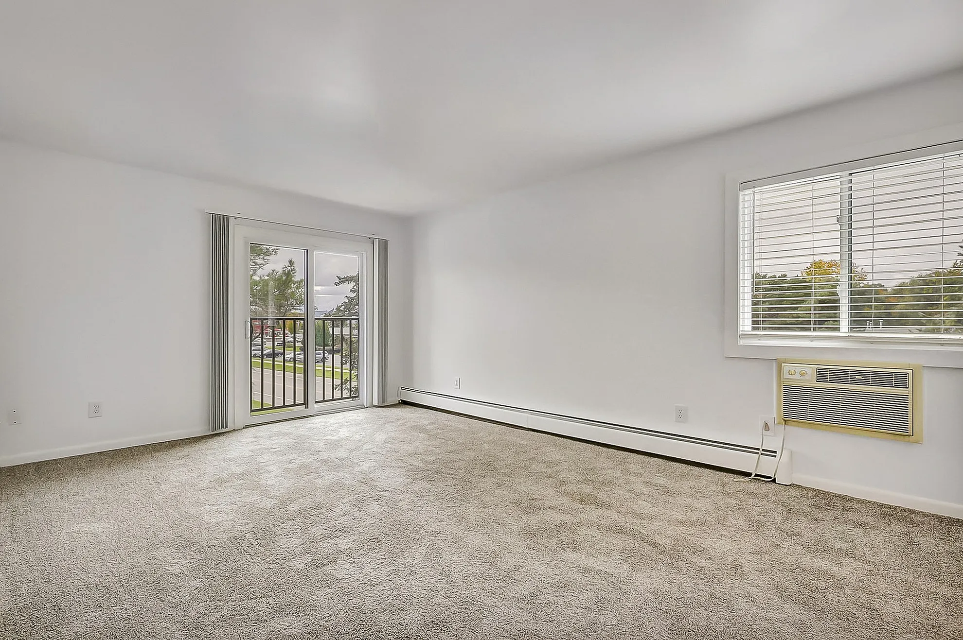 Bright living room with plush carpeting, large windows letting natural light flood in, and sliding glass doors leading to a cozy balcony with picturesque views.