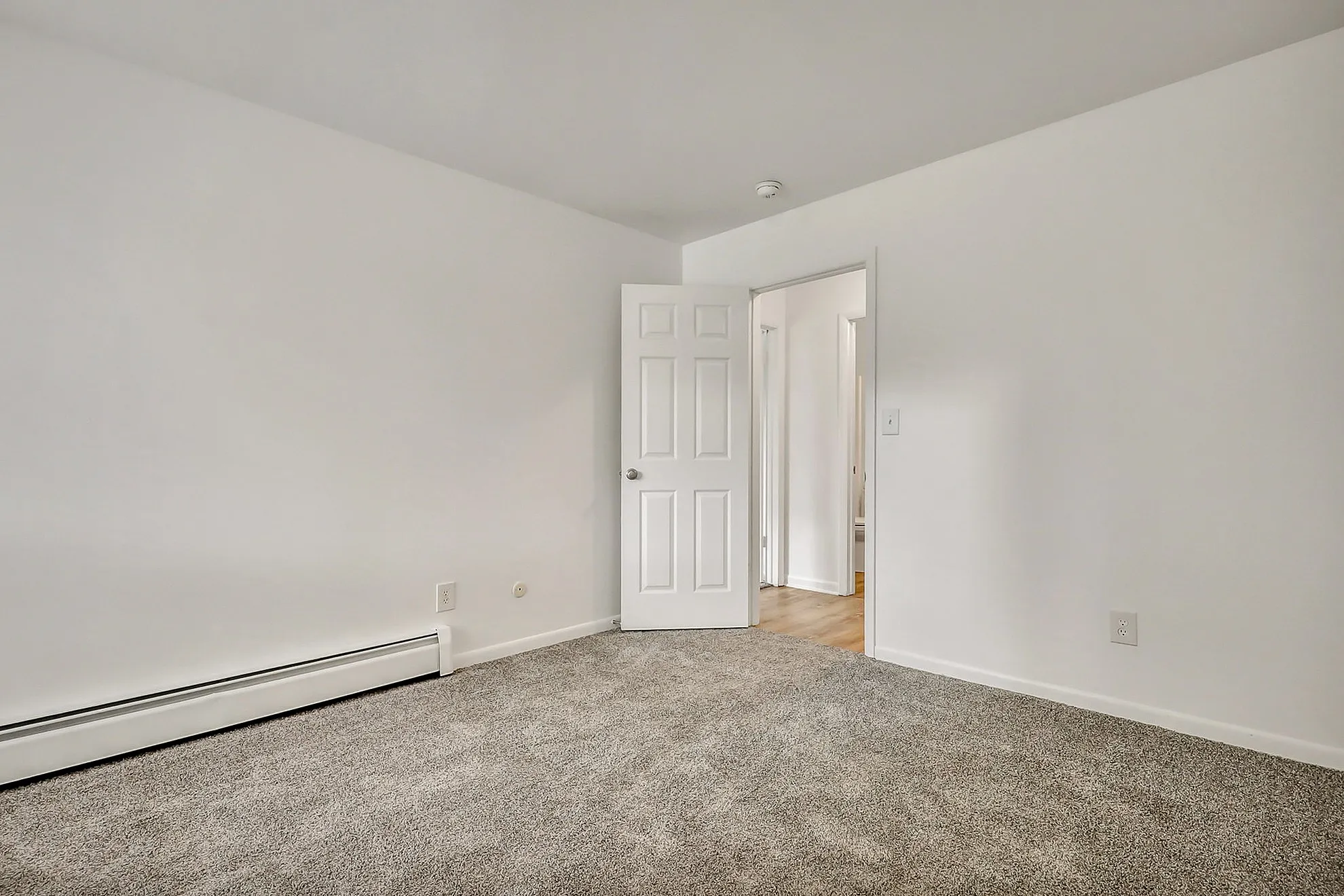 Bright, freshly painted room with plush carpeting and a white door leading to a warmly lit hallway.