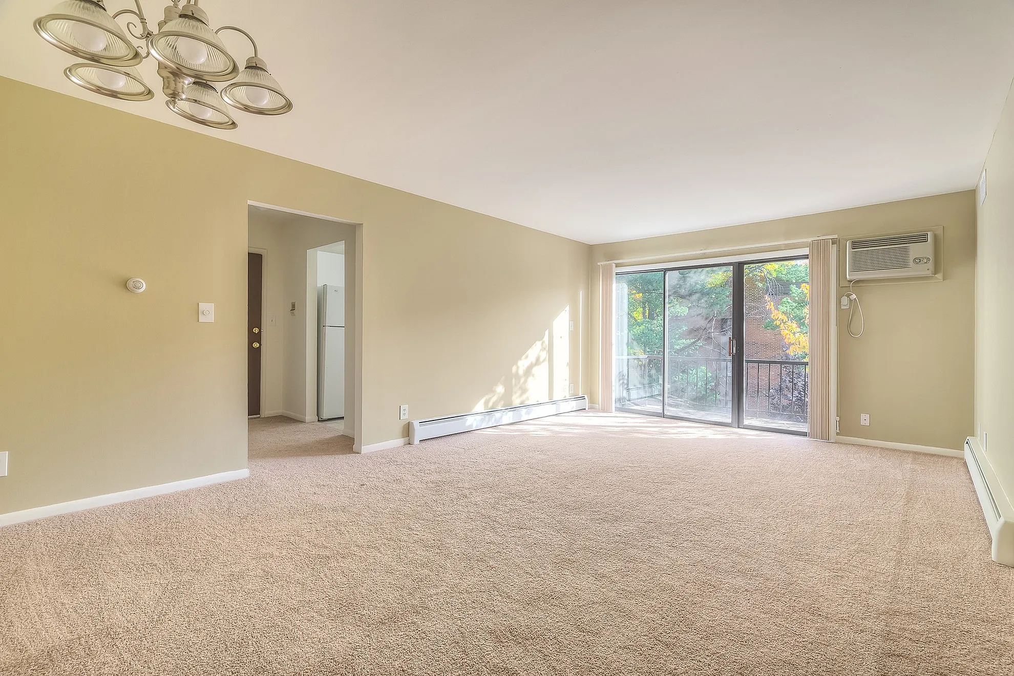 Bright and spacious living room with plush carpet, large sliding glass doors leading to a private balcony, and an elegant built-in air conditioning unit.