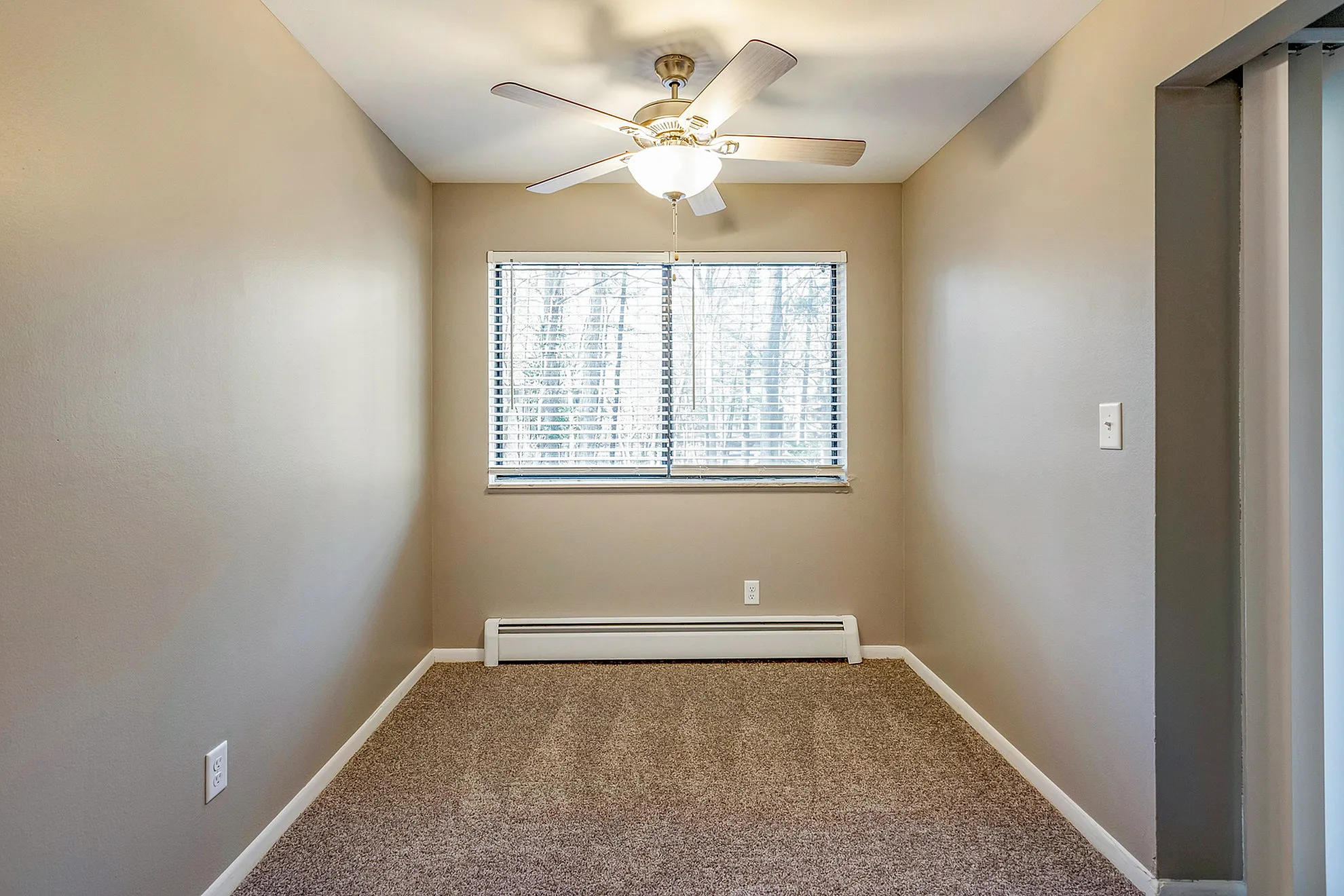Cozy room with elegant neutral walls, plush carpeting, and a large window allowing natural light, complete with a stylish ceiling fan.