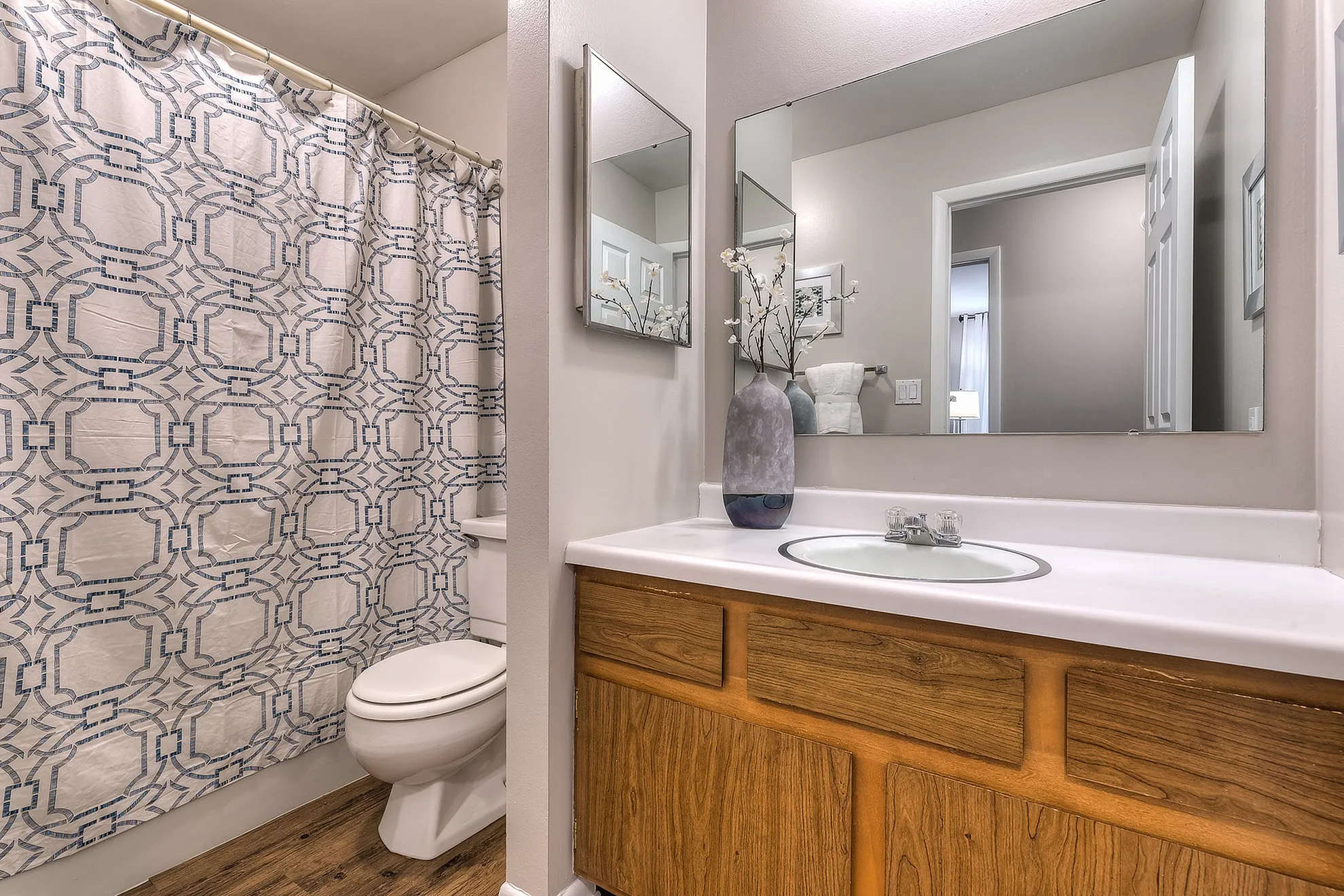 Bright and stylish bathroom featuring a modern sink, elegant mirror, and chic shower curtain, accentuated by natural wood cabinets and a decorative vase for a touch of elegance.